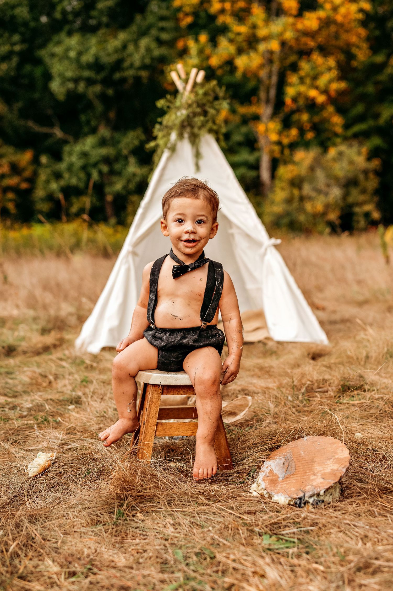 Toddler boy in black suspenders and bow tie sits on wooden stool outdoors near white teepee tent in autumn field.