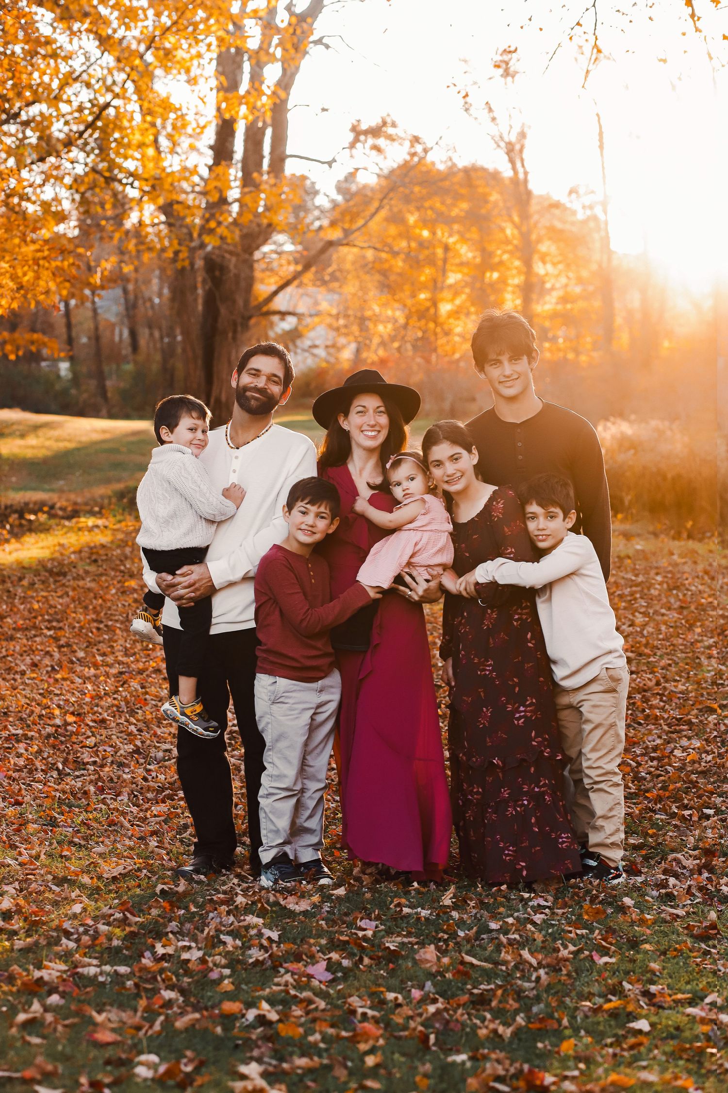 Family of eight posing outdoors among autumn foliage during golden hour sunset in a park setting.