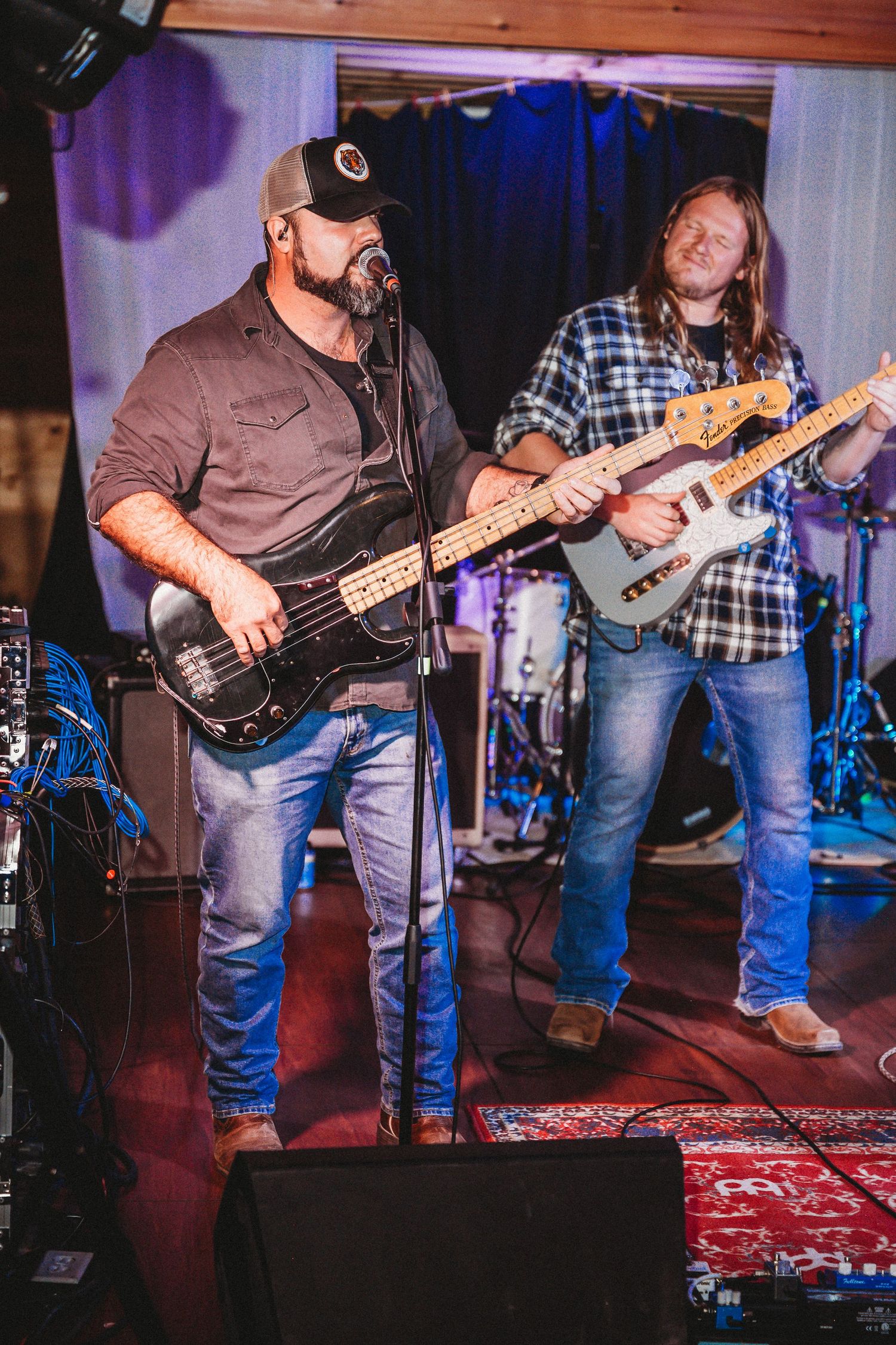 Two guitarists performing live on stage, one playing bass guitar wearing a cap, another in plaid shirt, at an indoor venue.