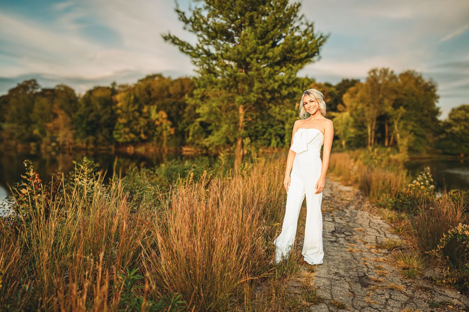 Beautiful blonde bride in a white jumpsuit stands on a scenic path surrounded by lush greenery and wildflowers.