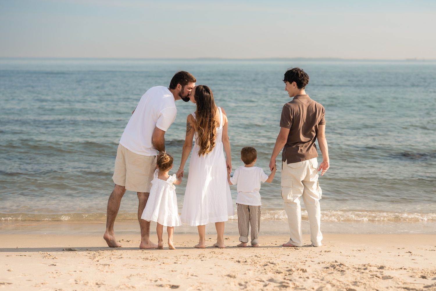 Family of five standing together on sandy beach looking at calm ocean on a sunny day.
