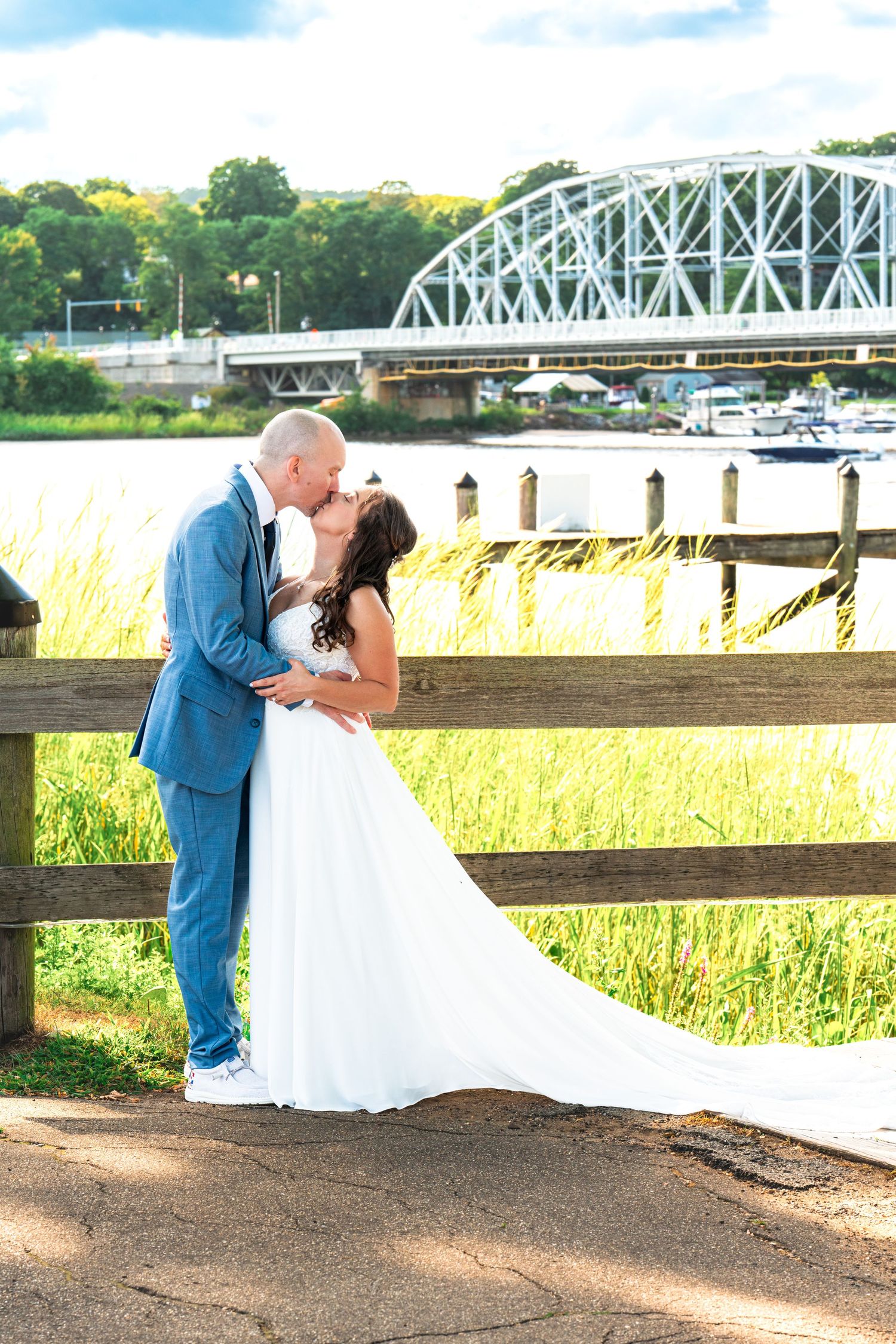 Bride and groom share a romantic kiss by a wooden fence near a river with a white bridge in the background.
