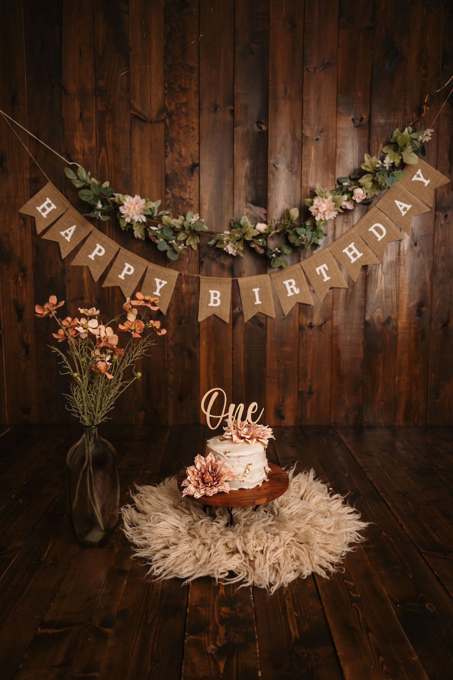 Rustic first birthday cake smash setup with burlap Happy Birthday banner, floral garland, and white cake with One topper.