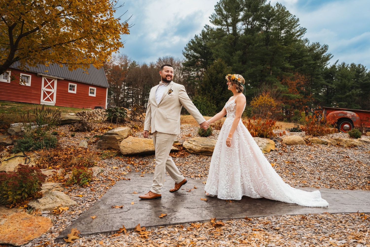 Bride and groom holding hands at a rustic fall wedding venue with a red barn and autumn foliage.