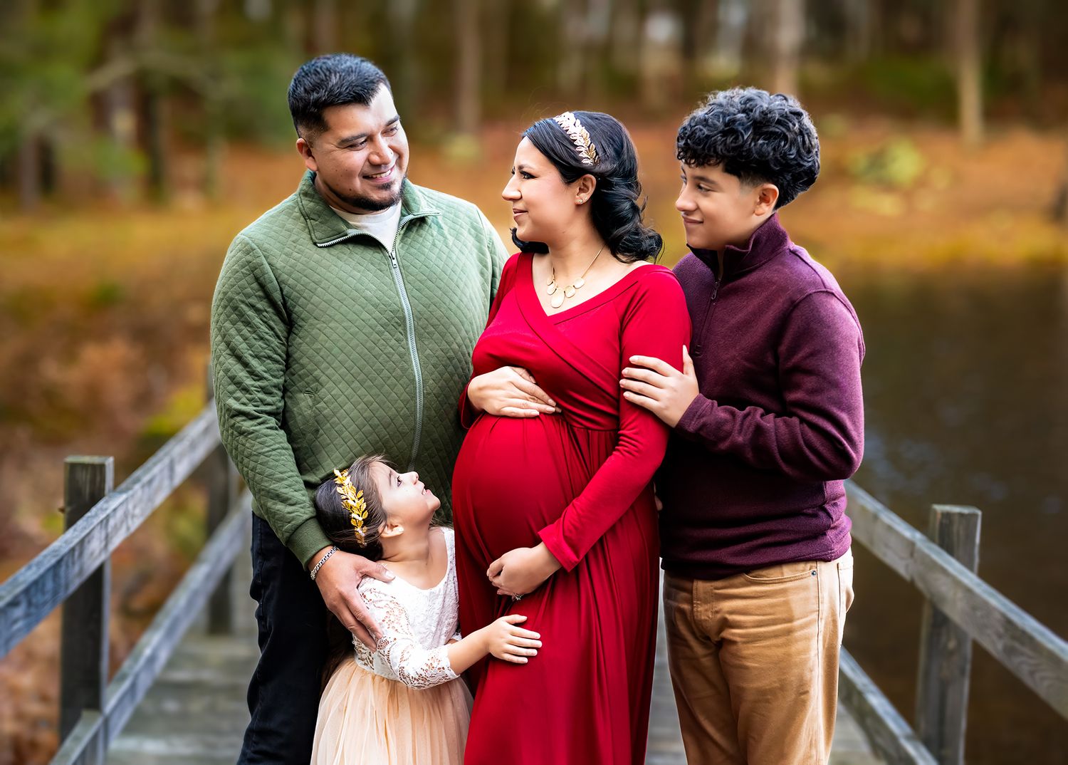 A joyful family of four poses on a wooden bridge outdoors, celebrating a maternity moment in autumn.