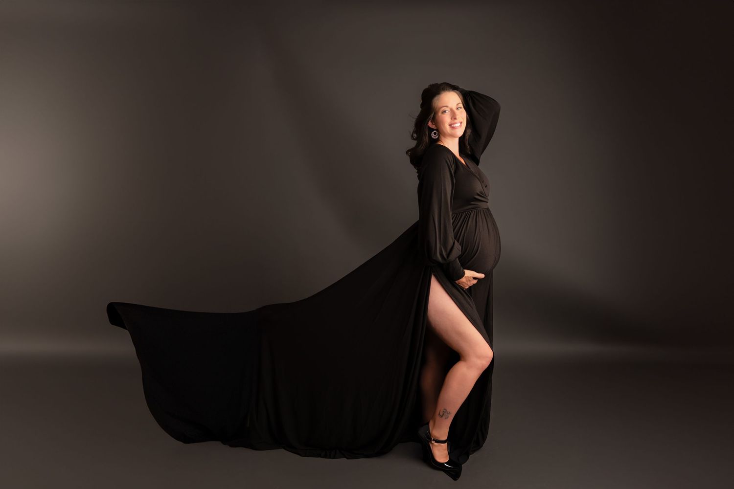 Pregnant woman in flowing black gown posing elegantly in studio, fabric trailing dramatically behind her.