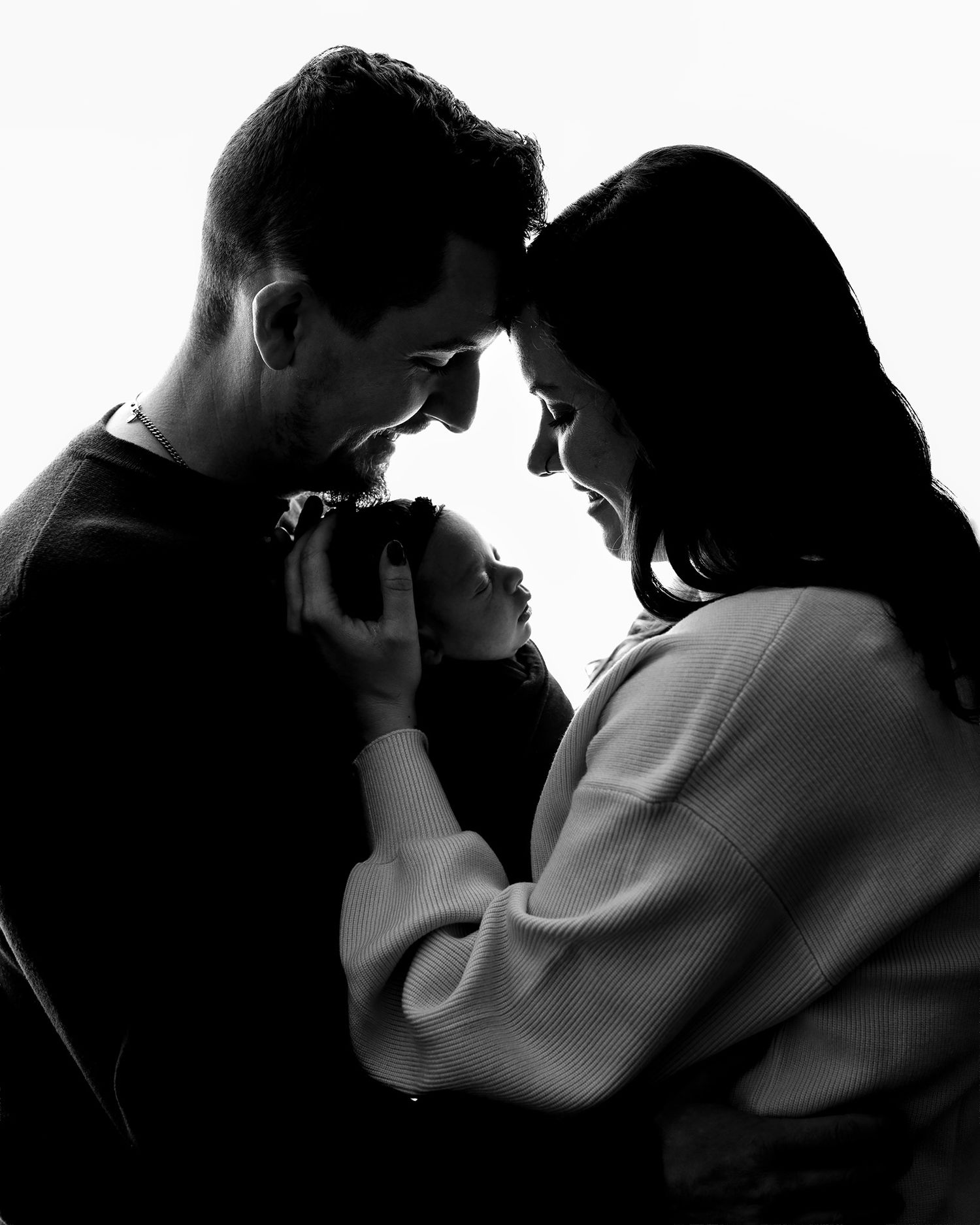 Black and white photo of two parents tenderly holding their newborn baby close together in an intimate family portrait.