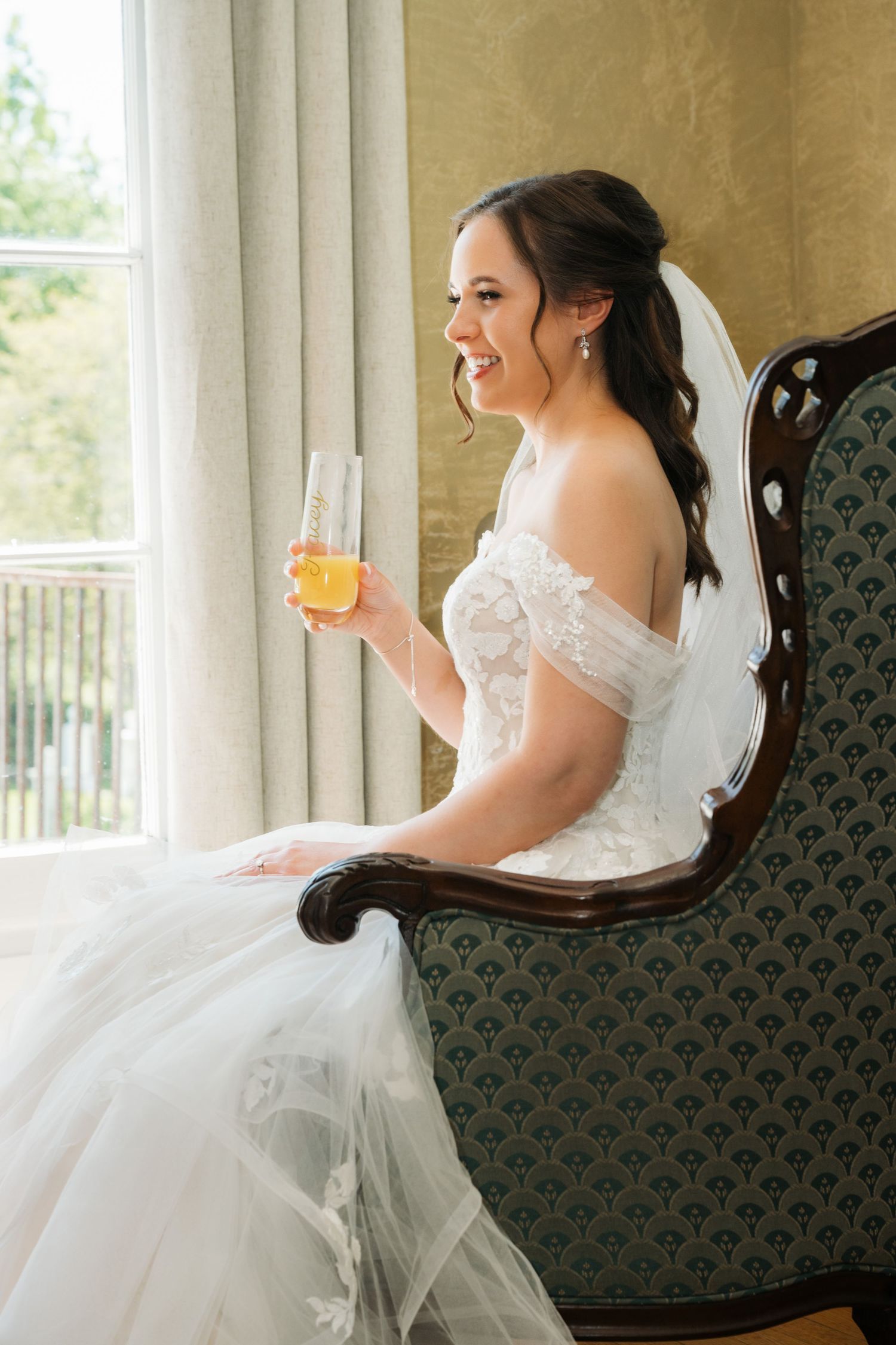 Smiling bride in off-shoulder lace gown holding champagne glass, seated on vintage chair by window on wedding day.