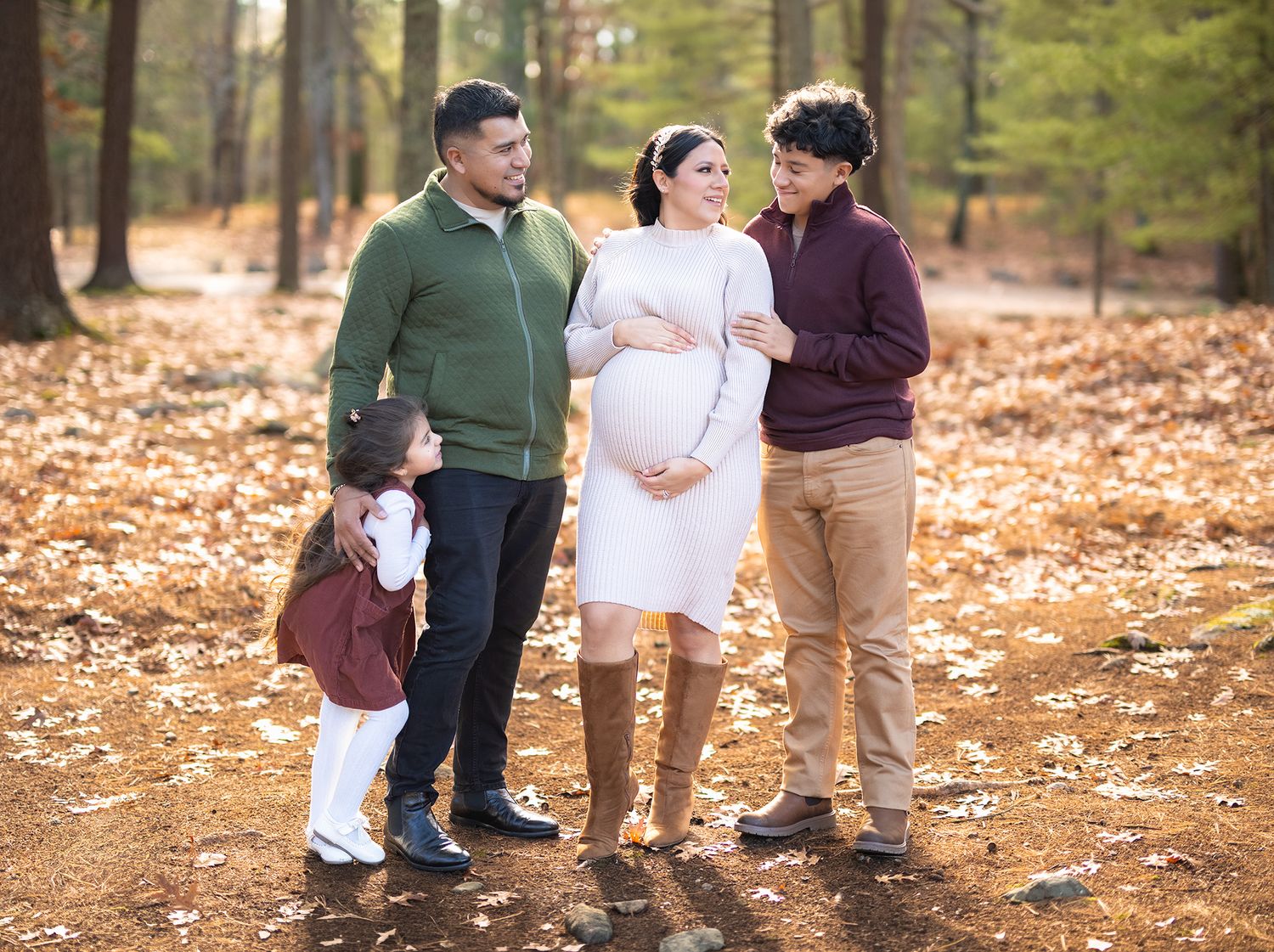 Pregnant woman in white dress poses with family in autumn forest setting for maternity photo shoot.