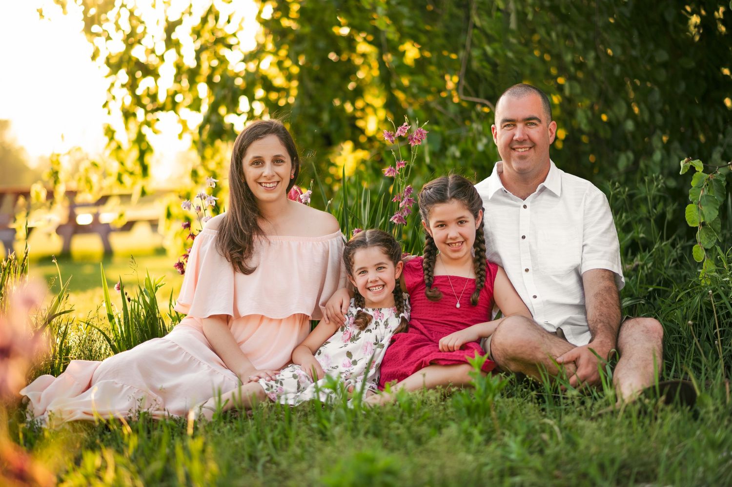 Family of four smiling outdoors in golden hour sunlight, sitting on green grass surrounded by lush garden greenery.