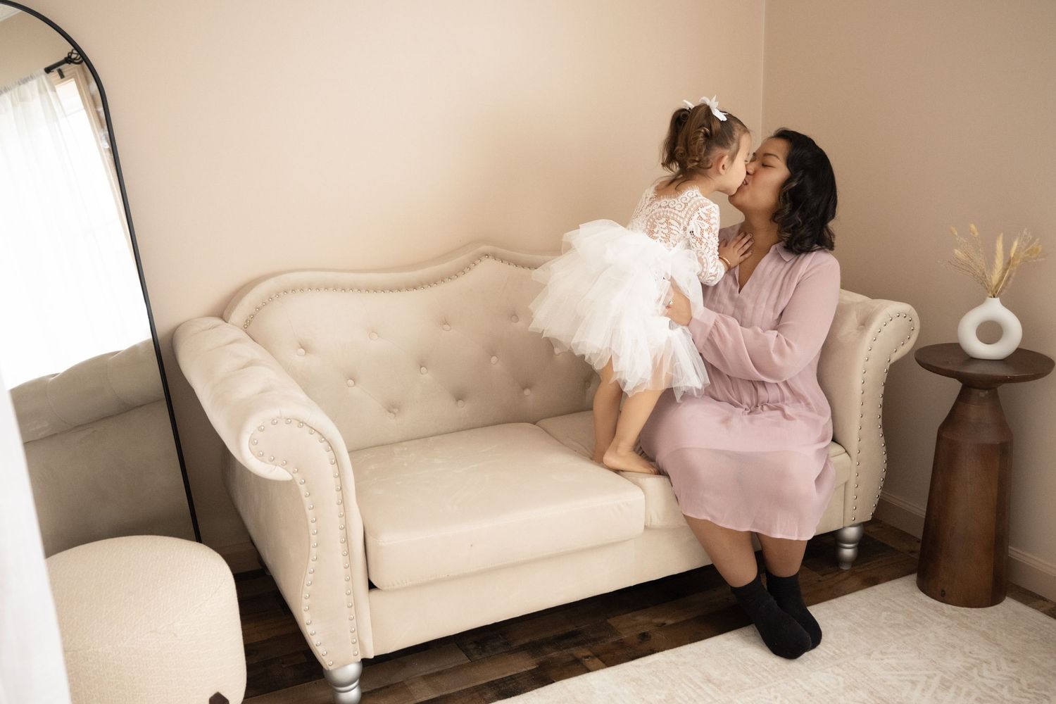 Loving mother shares a tender kiss with her young daughter in a white tutu dress on an elegant cream sofa.