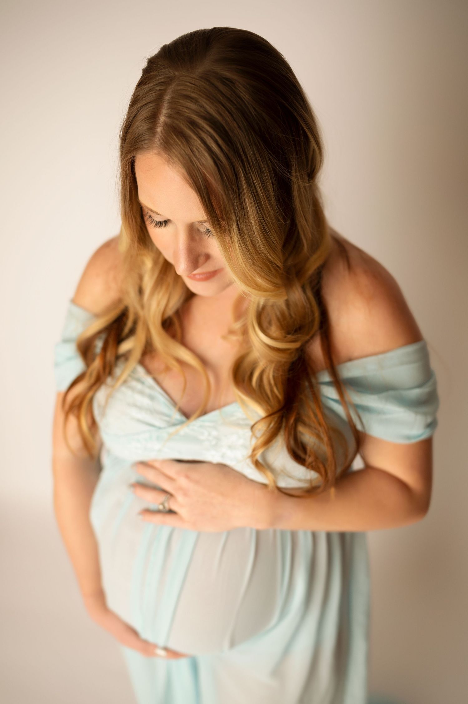 Pregnant woman in light blue off-shoulder gown cradling her baby bump, looking down tenderly in a soft studio setting.