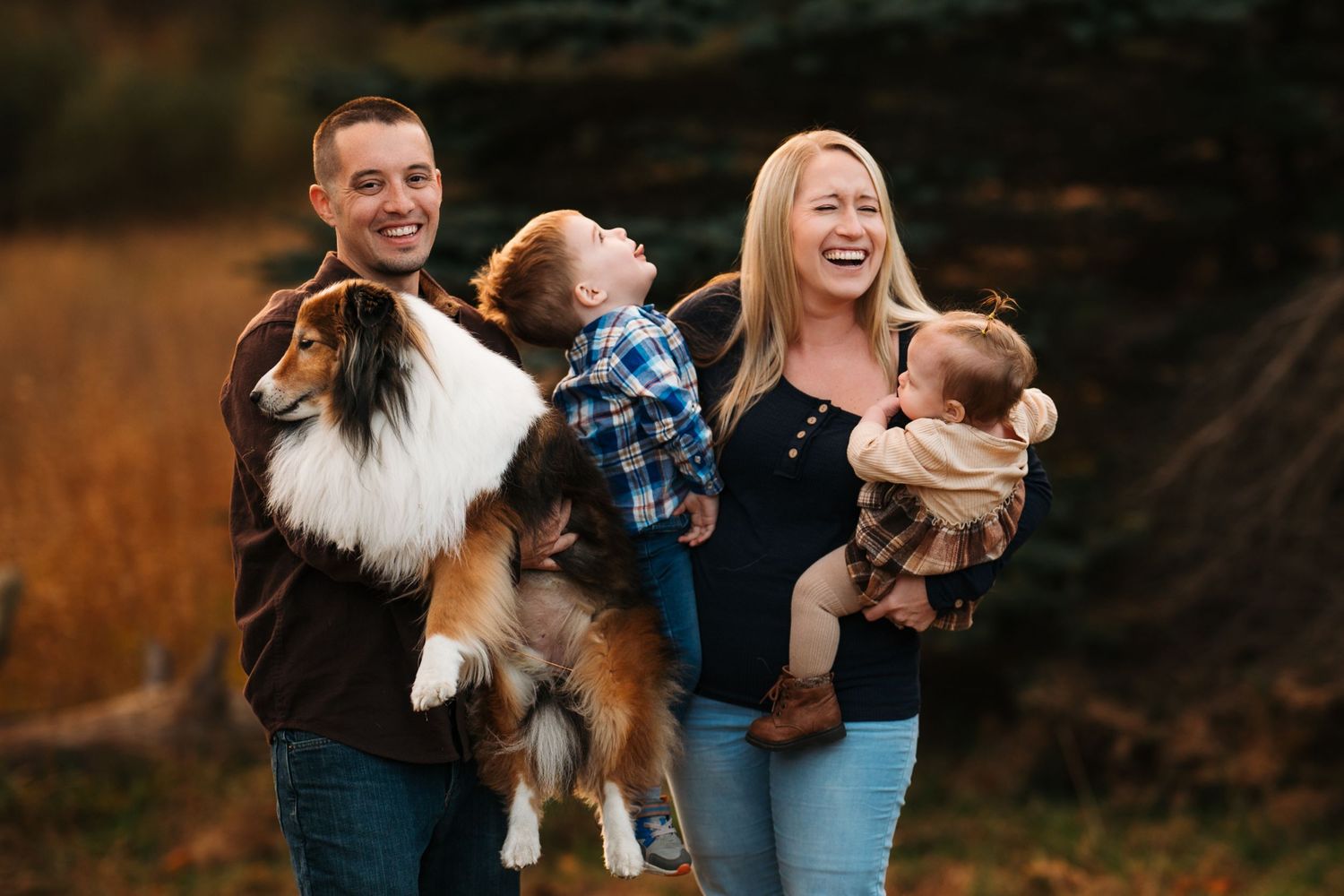 Happy family with two young children and a Sheltie dog laughing outdoors in a golden autumn field.