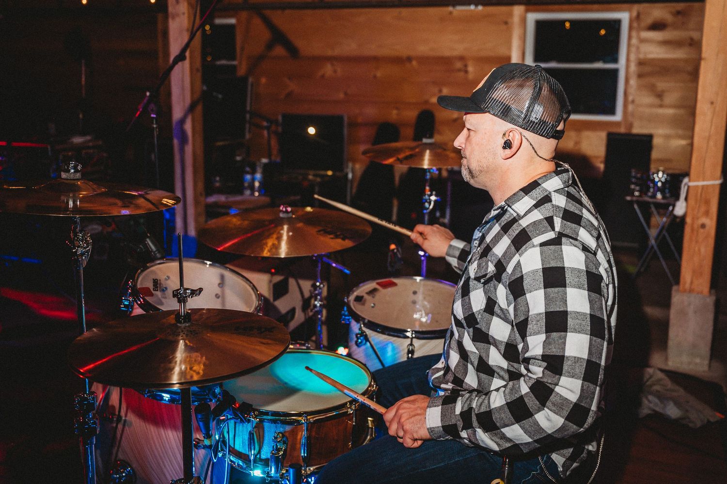 Drummer playing a blue drum kit on stage in a plaid shirt and cap at an indoor music venue.