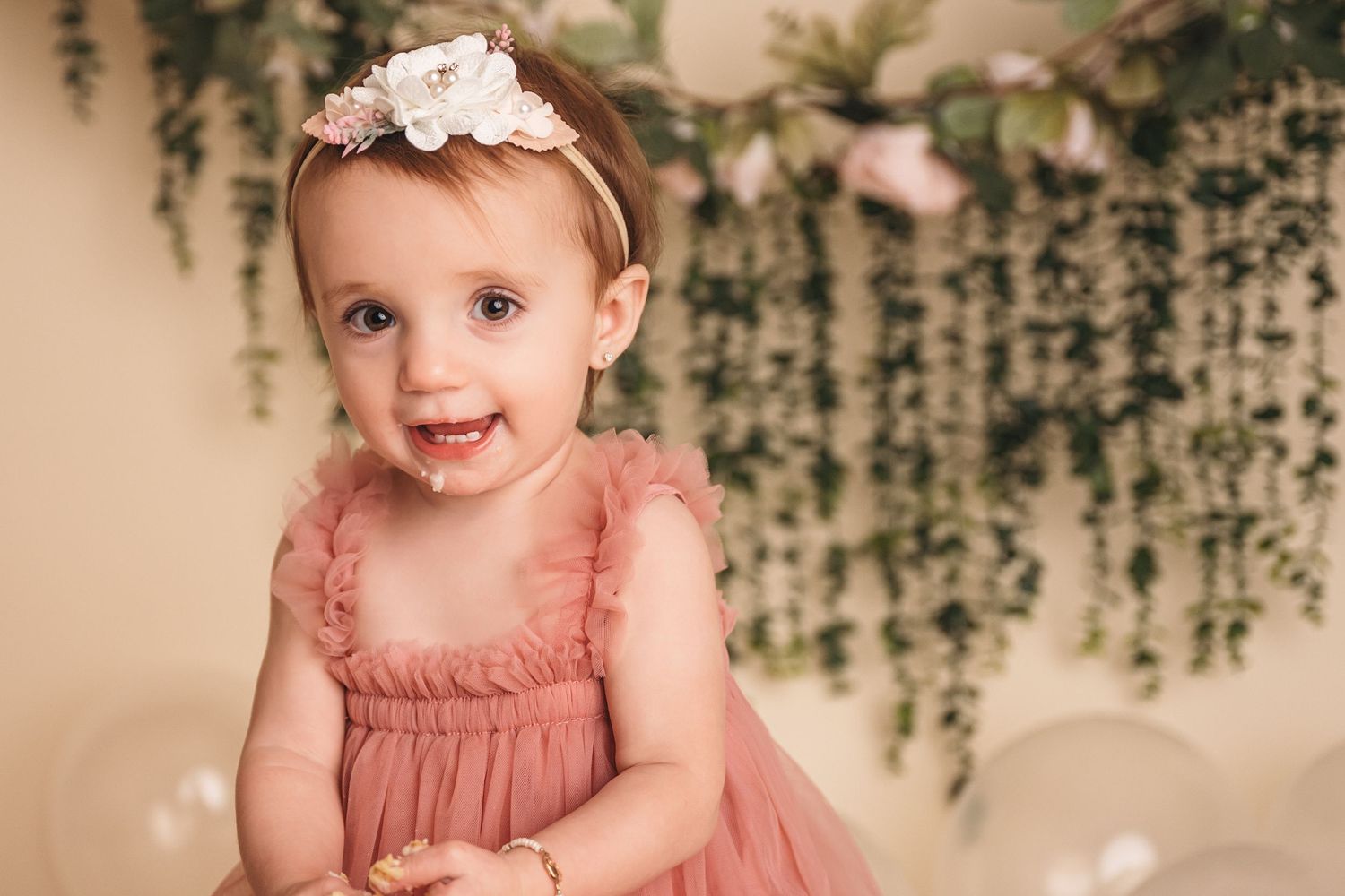 Smiling baby girl in pink tulle dress and floral headband at her first birthday cake smash photo session.