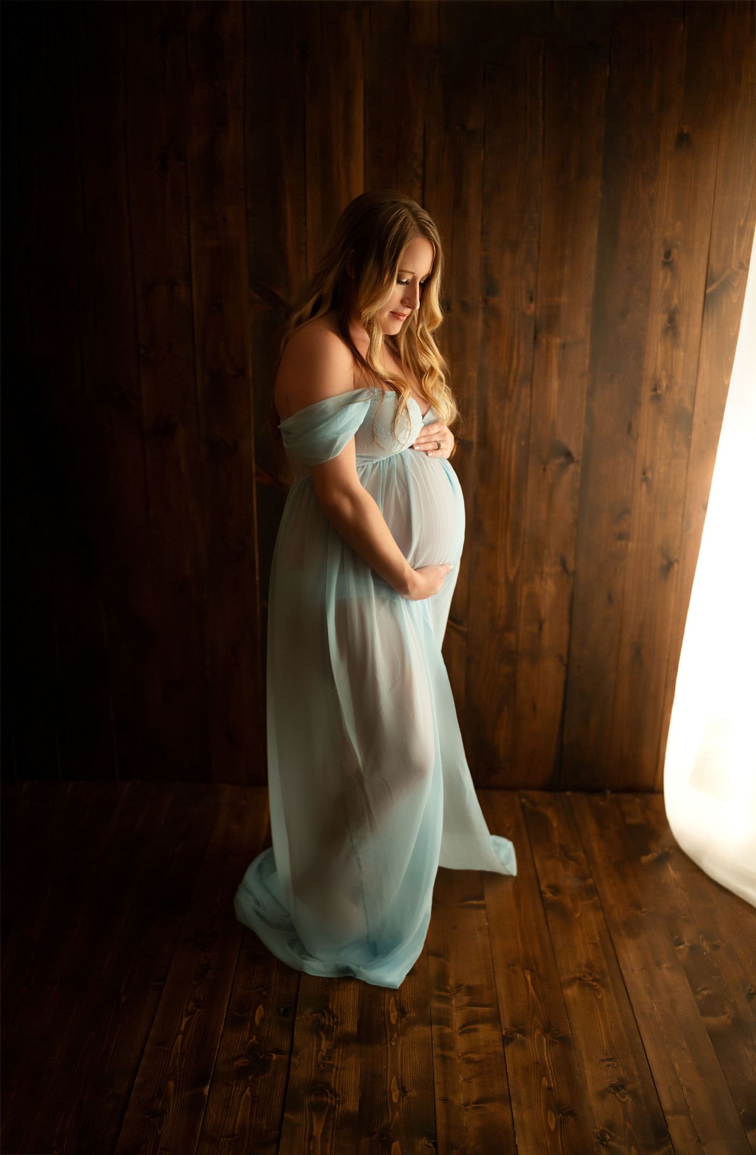 Pregnant woman in flowing light blue off-shoulder gown cradling baby bump, standing in warmly lit wooden studio.
