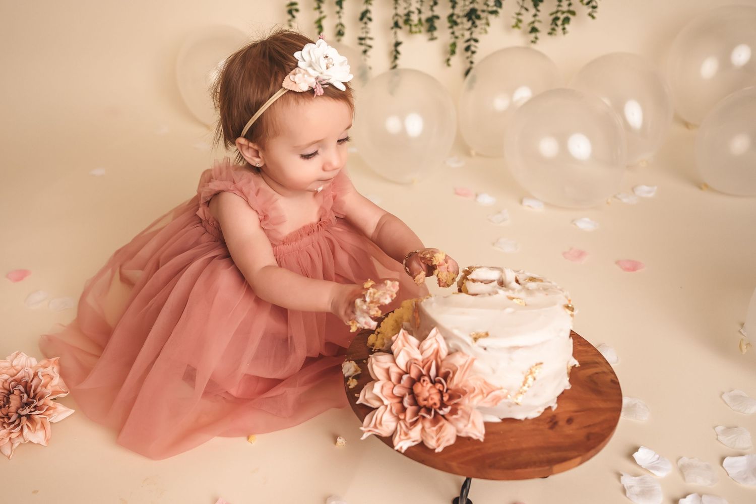 Baby girl in pink tulle dress touching a floral white cake during a first birthday smash cake photo session.