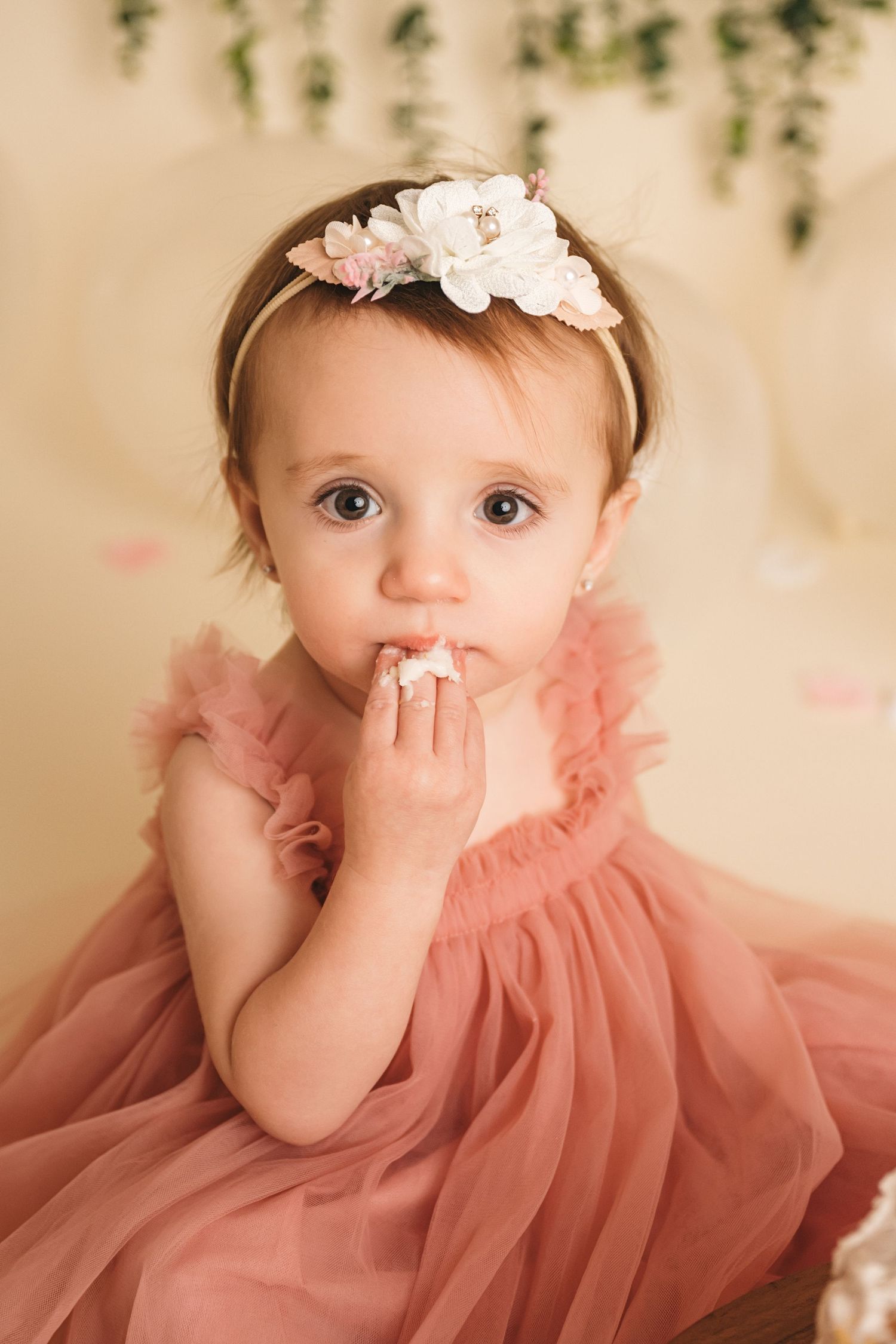 Adorable baby girl in pink tulle dress and floral headband eating cake during first birthday smash cake photoshoot.