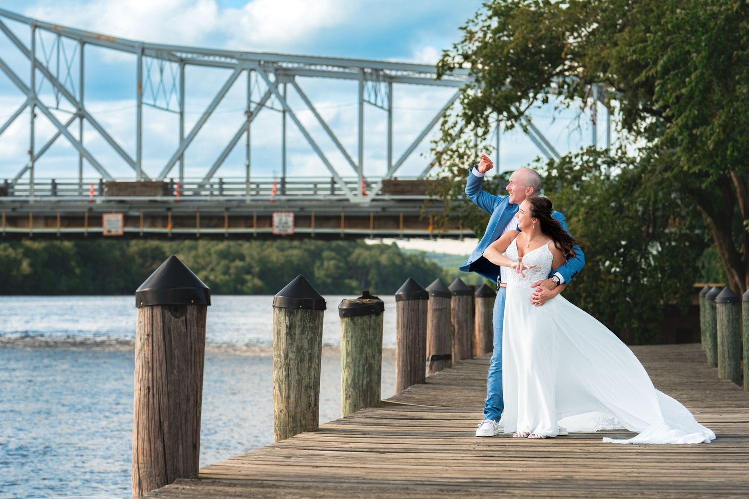 Bride and groom pose romantically on a waterfront dock with a steel bridge backdrop on their wedding day.