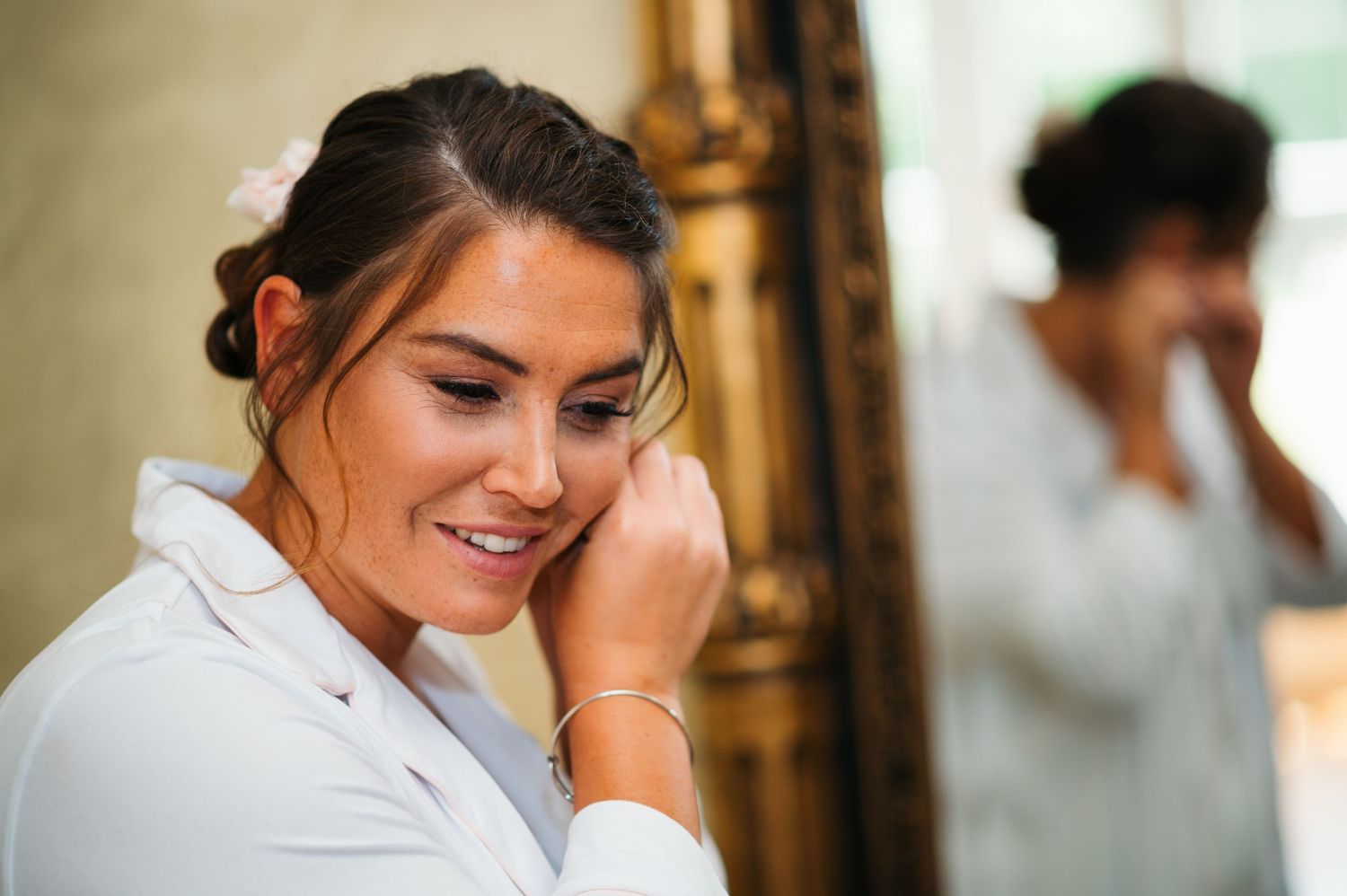Bride in white robe smiling while putting on earrings, with reflection visible in ornate gold mirror.