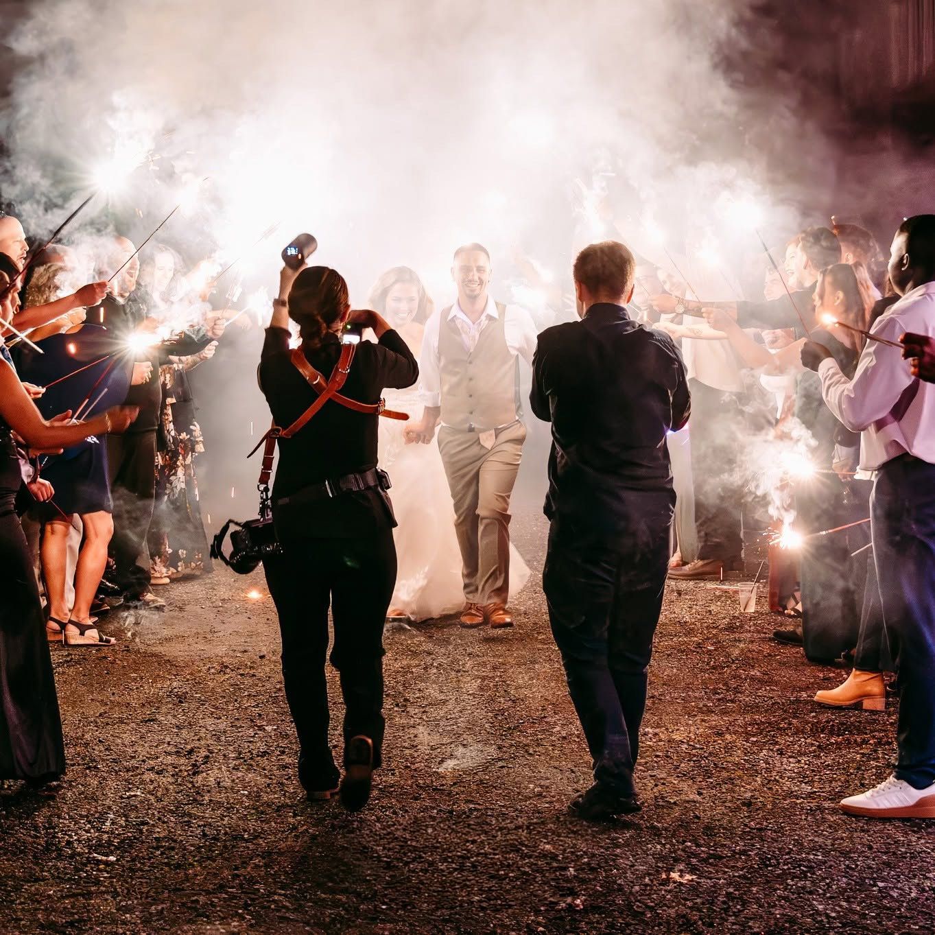 Bride and groom walk through a sparkler tunnel surrounded by cheering guests at a romantic nighttime wedding exit.