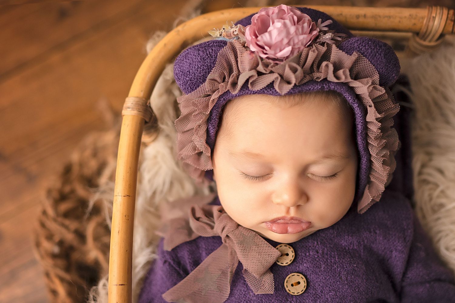 Sleeping newborn baby in purple knit outfit and floral bonnet resting in a wicker basket on a wooden floor.