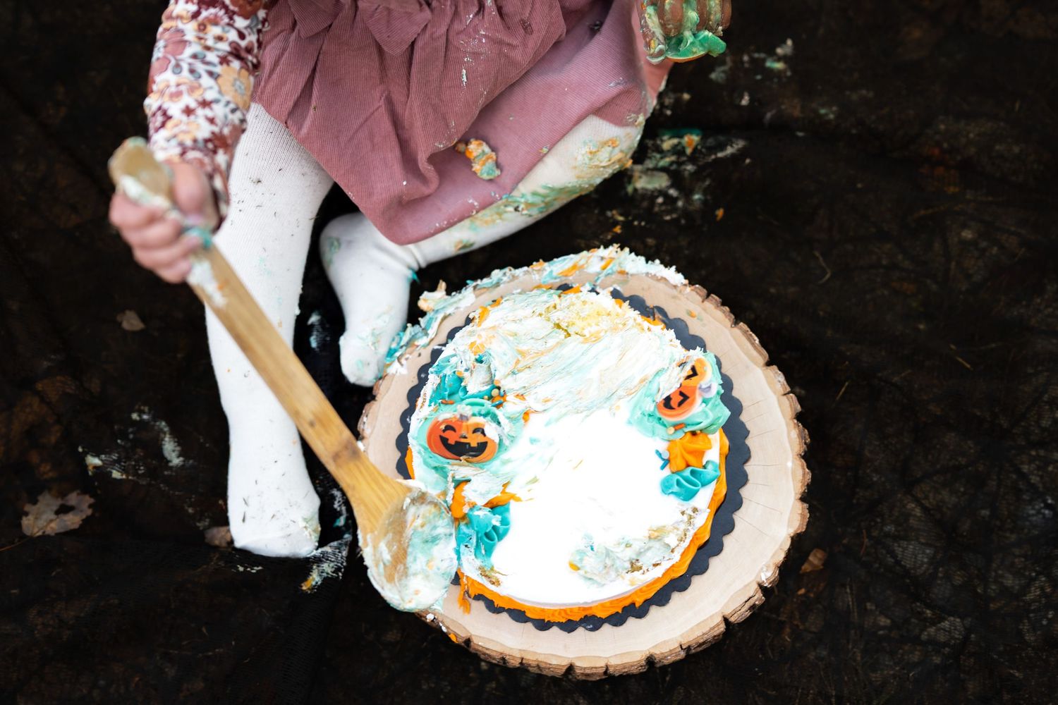Child stirring a decorated white cake with a wooden spoon outdoors on dark ground.