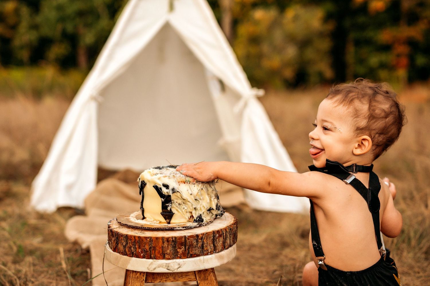 Happy toddler in black romper reaches for smash cake on wood stump with white teepee backdrop outdoors.