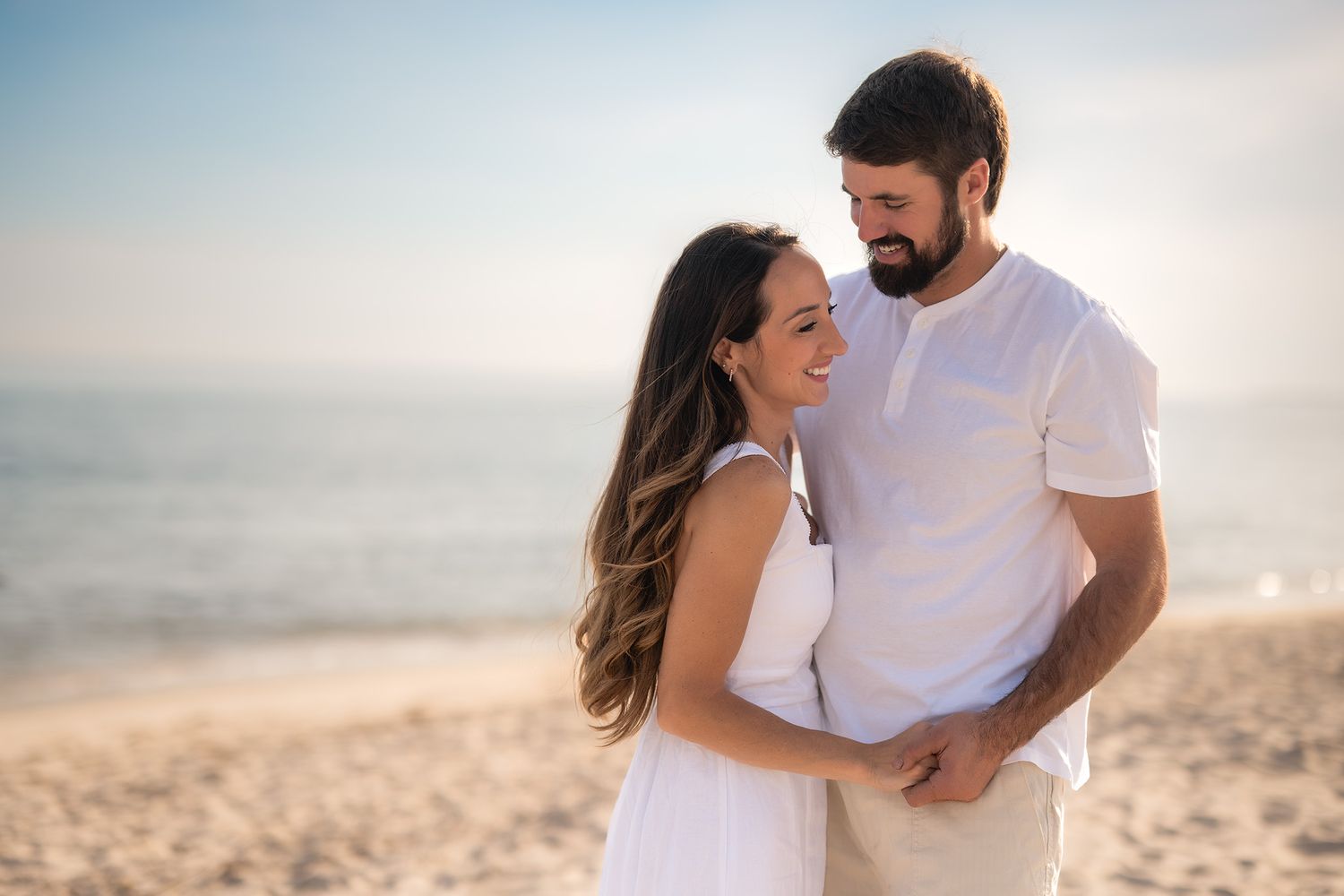 Romantic couple in white outfits embracing on a sandy beach at golden hour sunset.