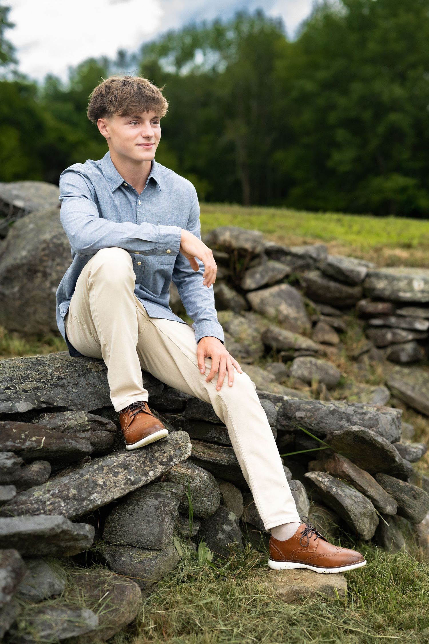 Young man in blue shirt and khaki pants sitting on a stone wall in a scenic outdoor setting.
