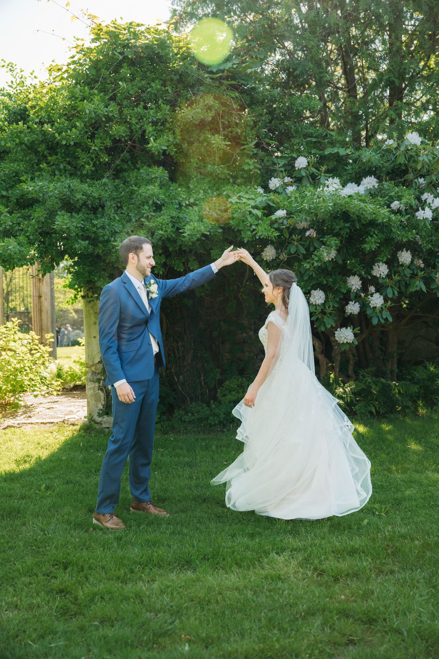 Groom in blue suit twirling bride in white gown outdoors on green lawn with lush garden backdrop.