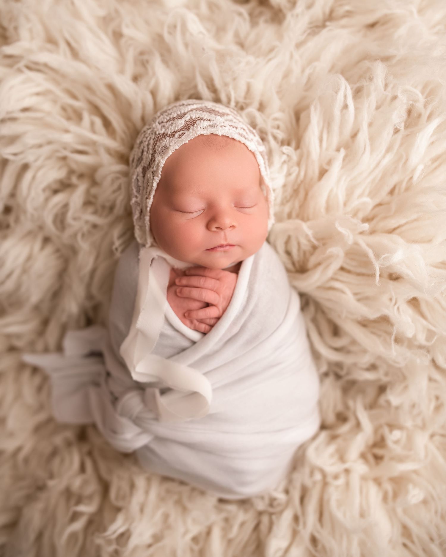 Sleeping newborn baby swaddled in white wrap wearing lace bonnet on fluffy cream fur rug.