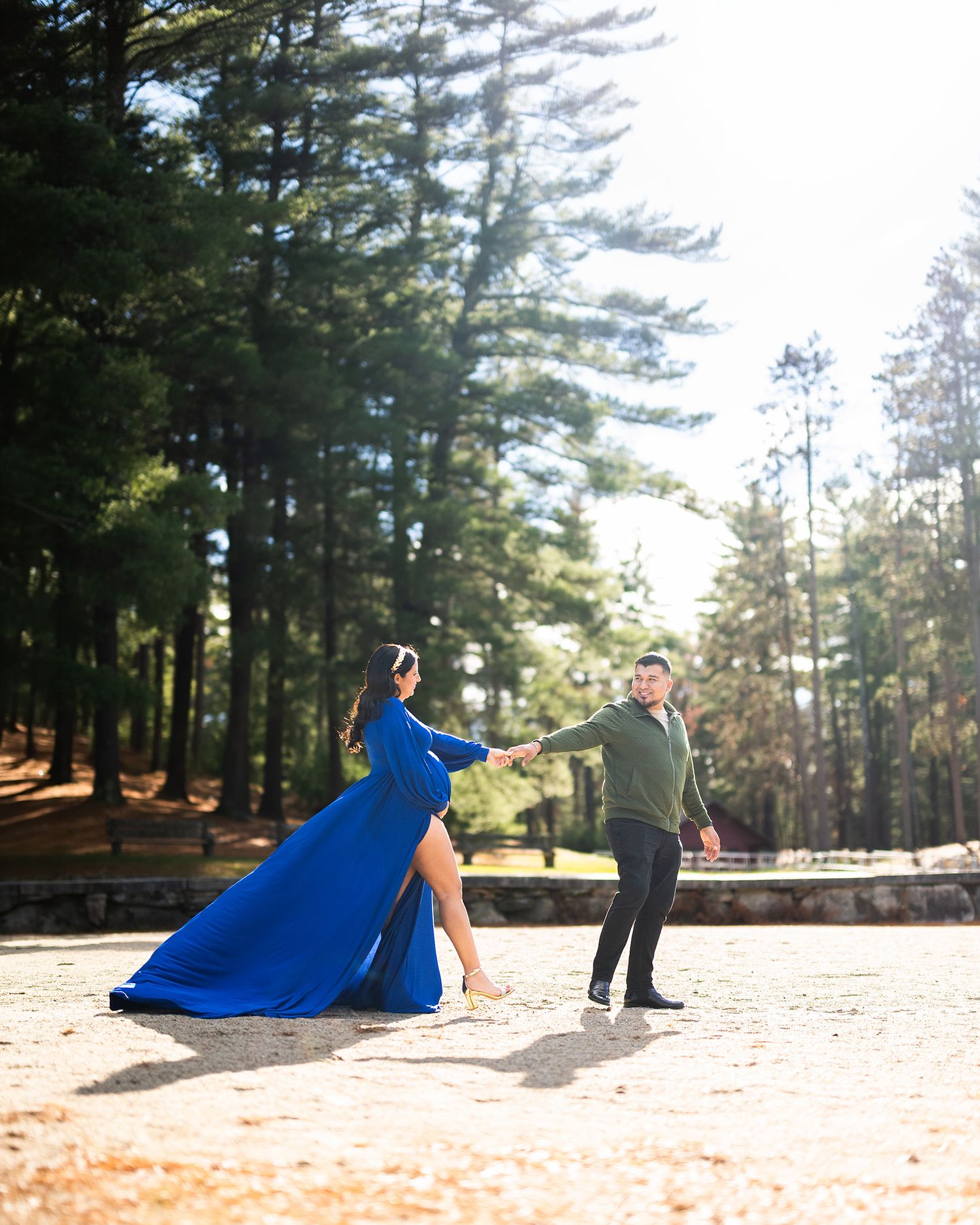Couple dancing outdoors, woman in flowing blue gown, man in green sweater, surrounded by tall pine trees.