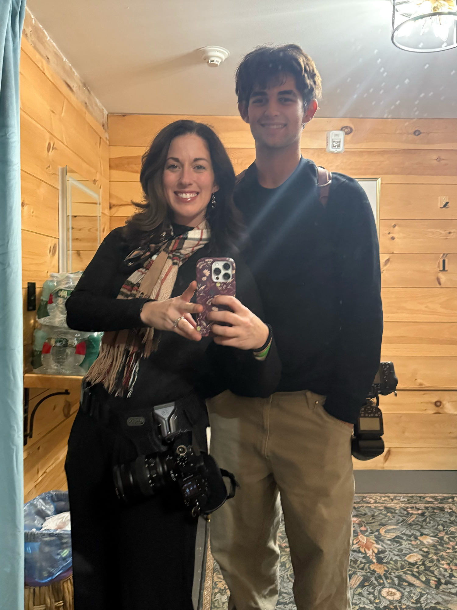 Two photographers smile in a mirror selfie inside a wood-paneled cabin, both wearing cameras and casual outfits.