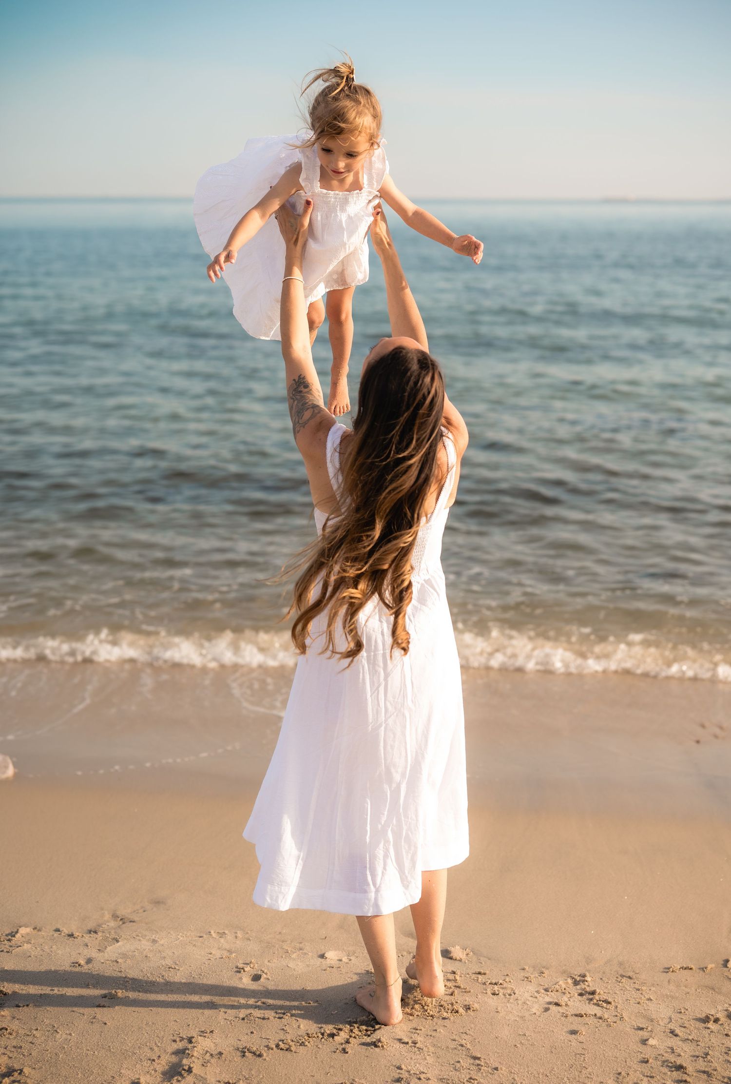 A mother in a white dress lifts her young daughter in the air on a sunny beach by the ocean.