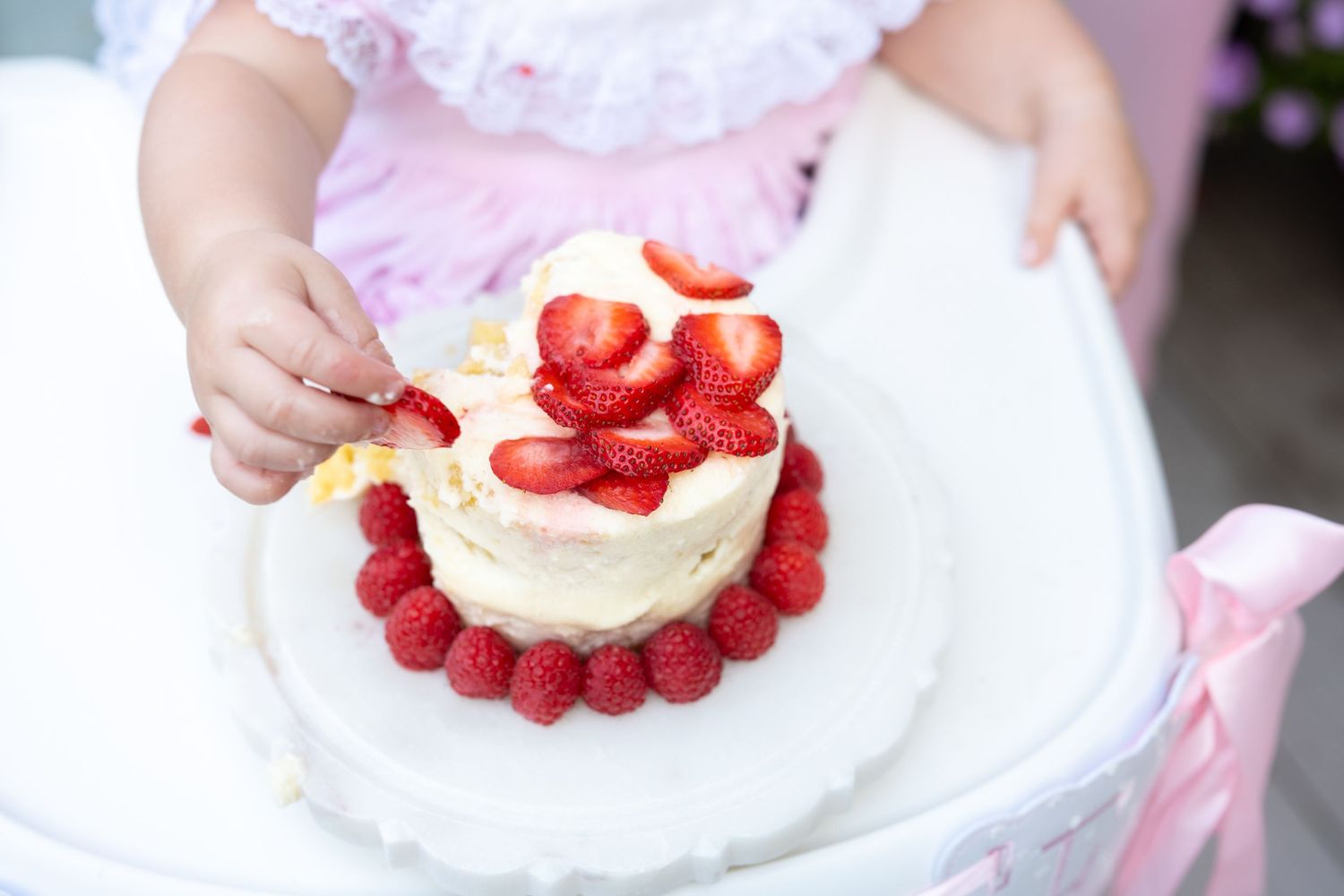 Baby girl in pink dress reaching into a white cake topped with fresh strawberries and raspberries for first birthday.