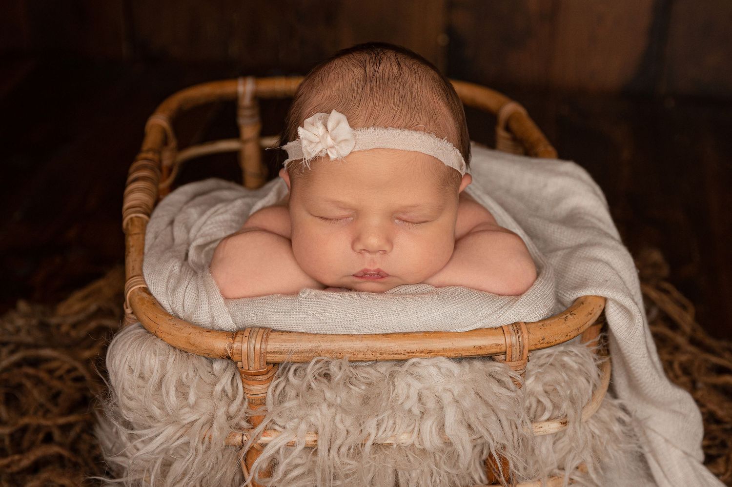 Sleeping newborn baby in a wicker basket with a white floral headband on a fluffy gray blanket.