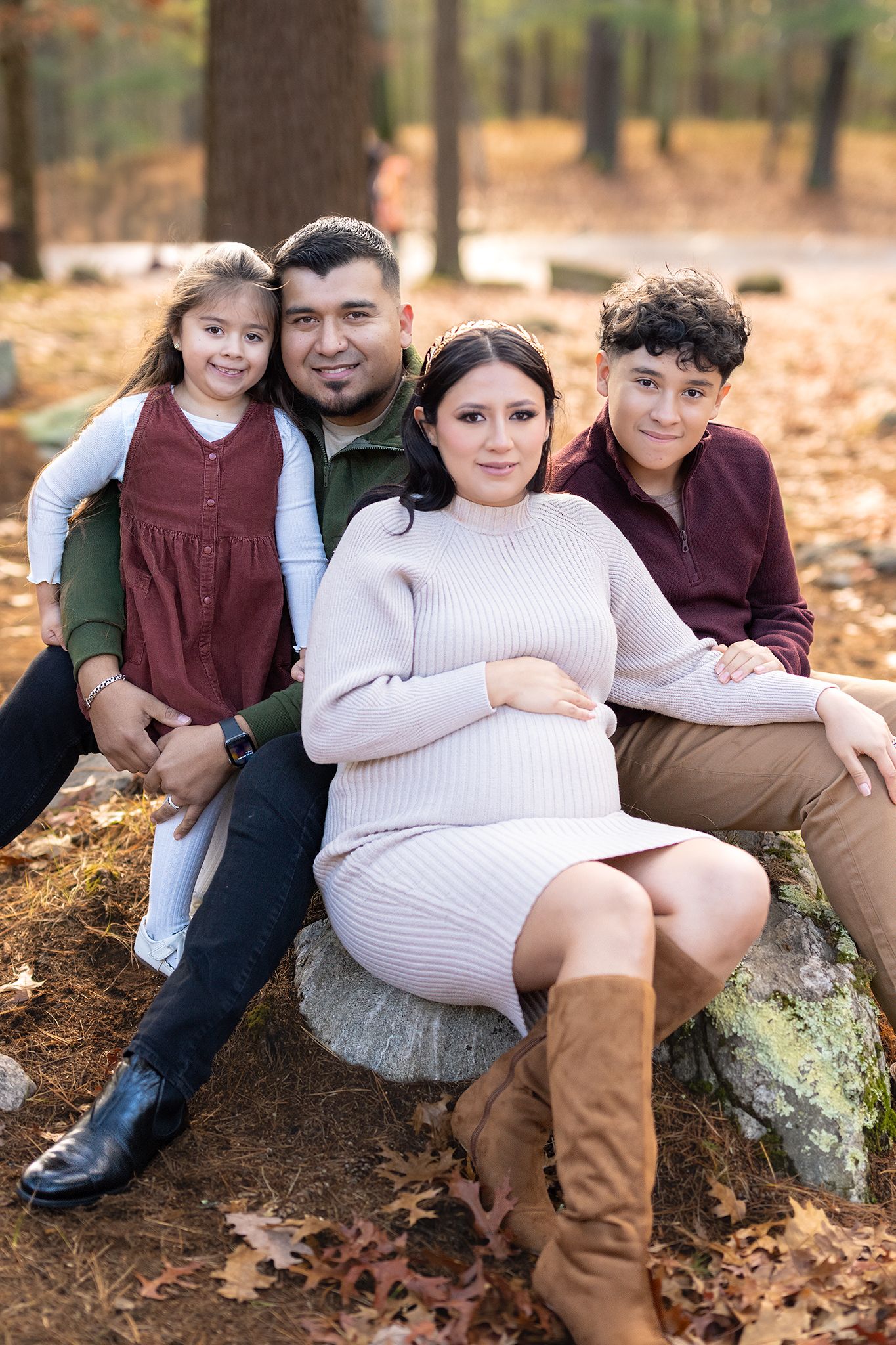 Happy Hispanic family with pregnant mother sitting on rocks during autumn forest photo session.