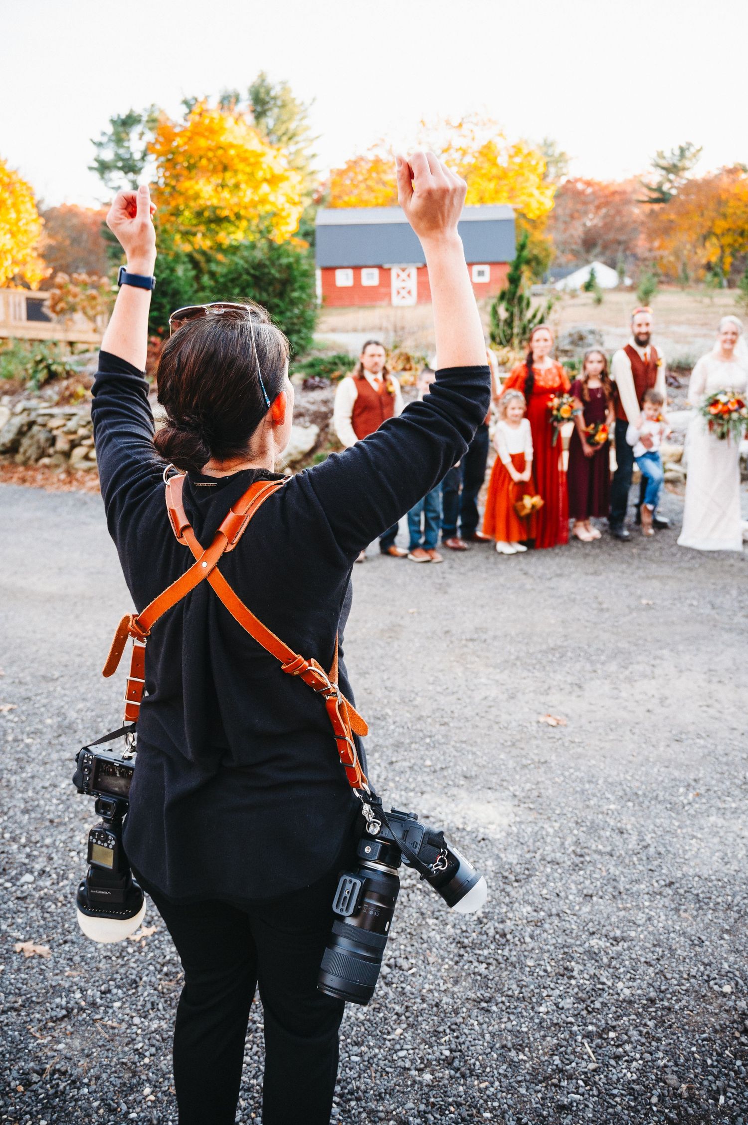 Wedding photographer with cameras raises hands directing bridal party outdoors at fall venue with red barn.