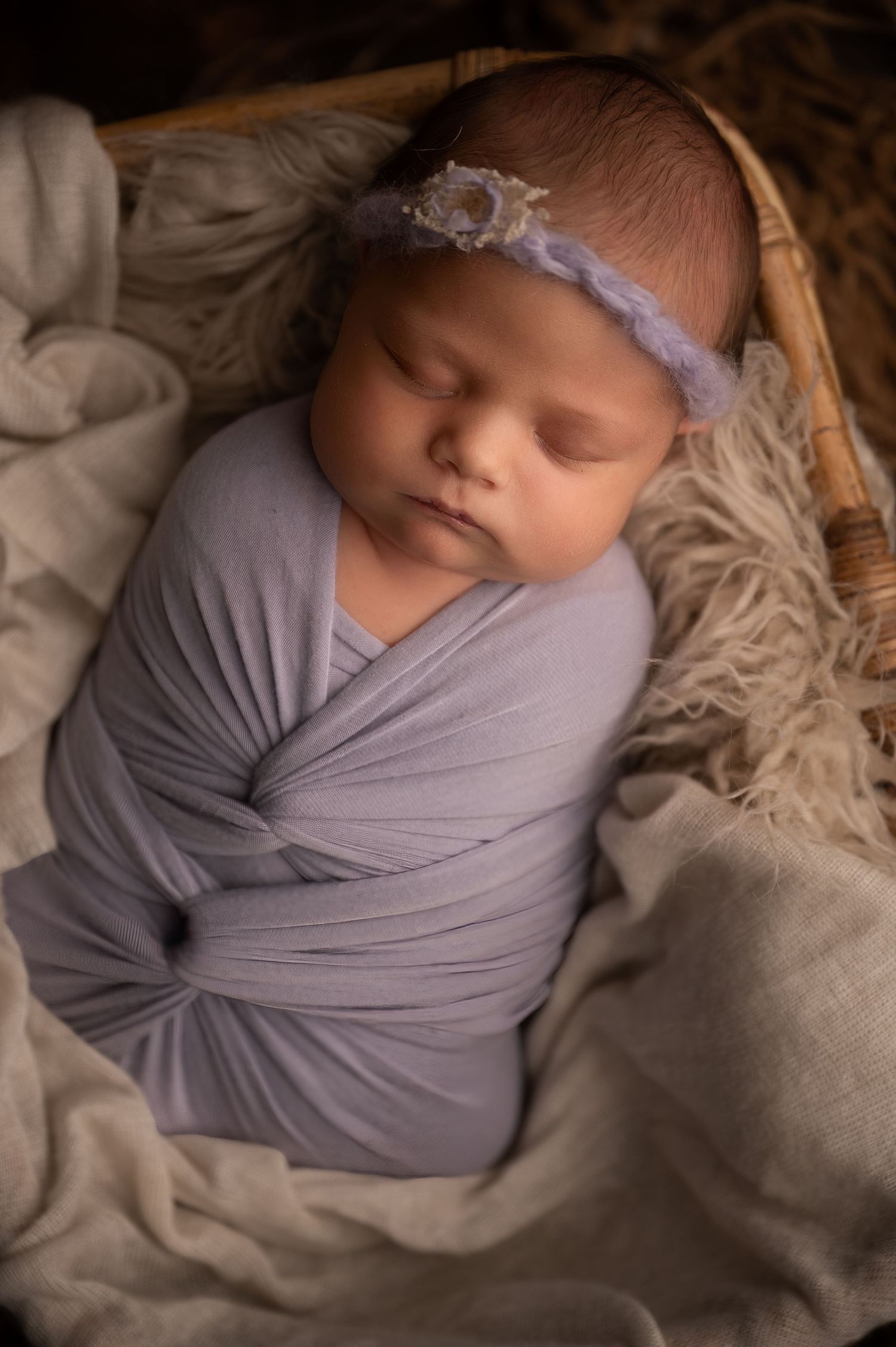 Newborn baby girl swaddled in lavender wrap with floral headband sleeping peacefully in a wicker basket.