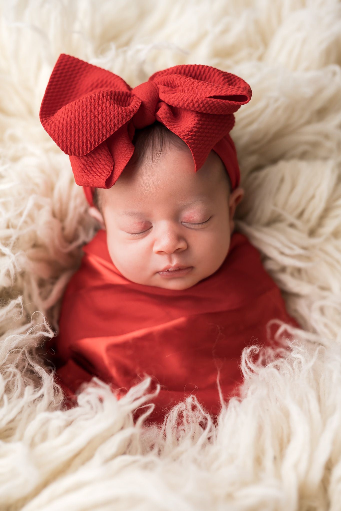 Sleeping newborn baby girl swaddled in red wrap with large red bow headband on fluffy cream fur backdrop.