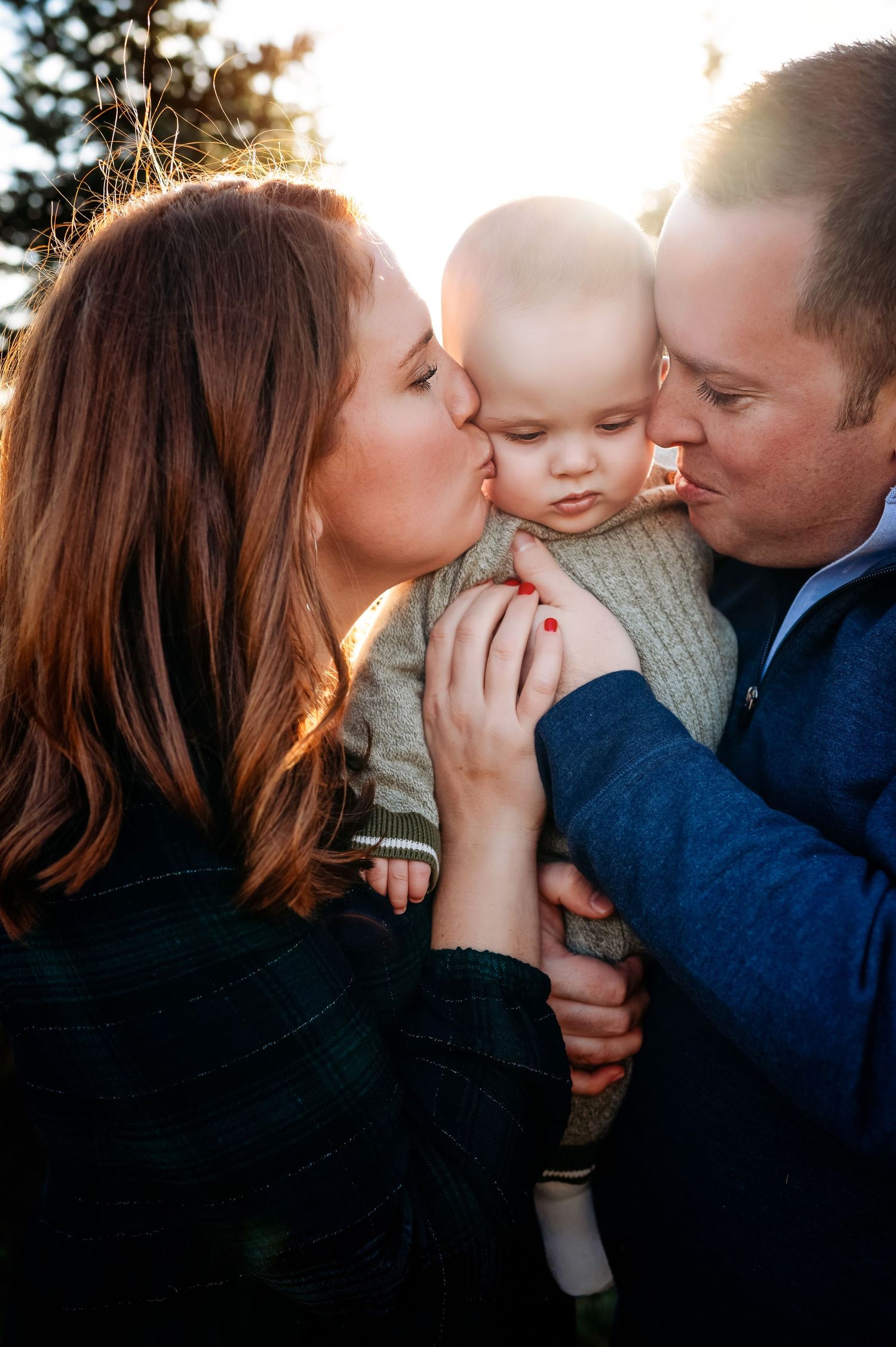 Parents kissing their baby on the cheeks in a warm golden sunset outdoor family portrait photo.