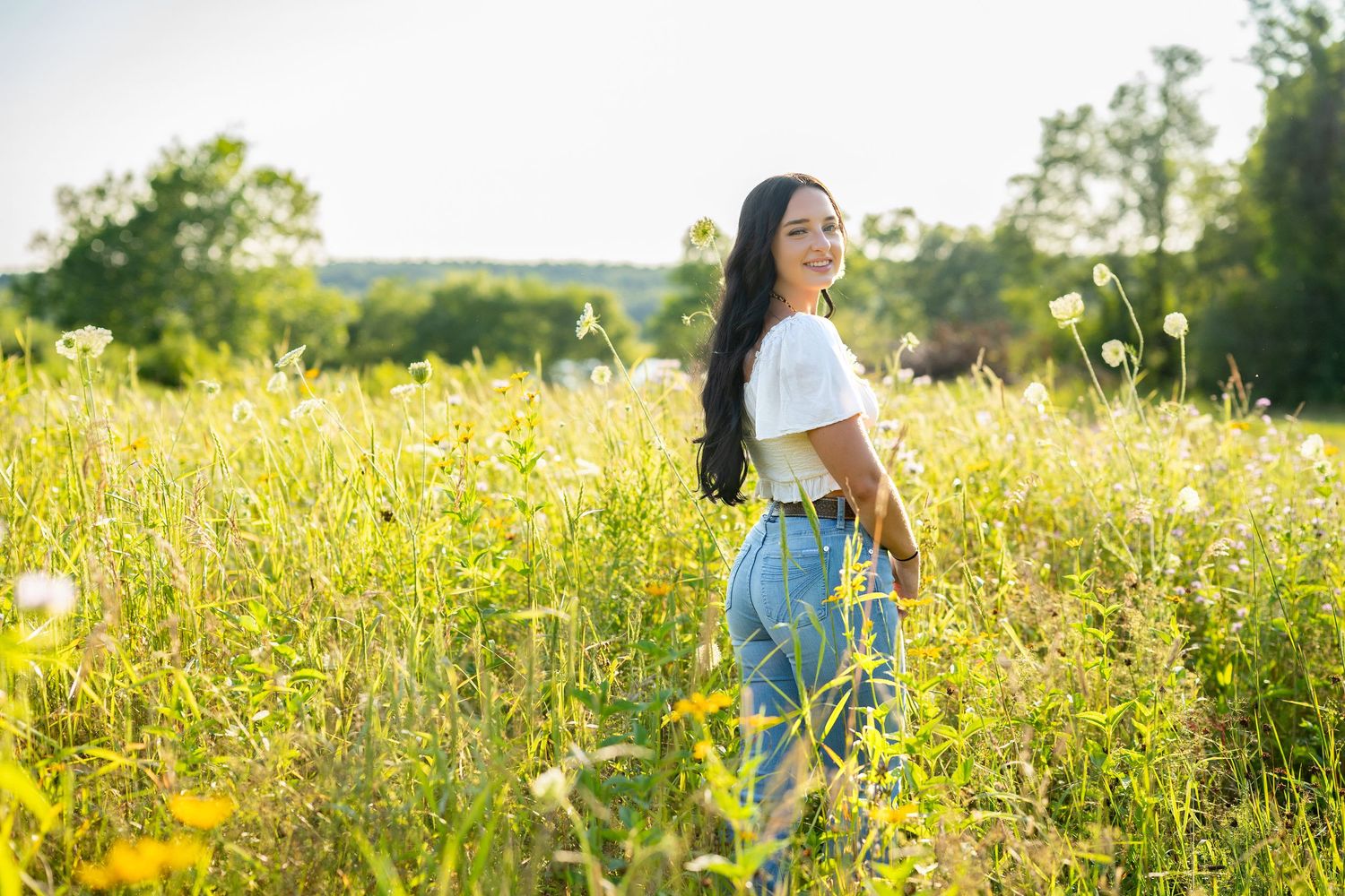 Young woman with long dark hair in white top and jeans standing in a sunny wildflower field.