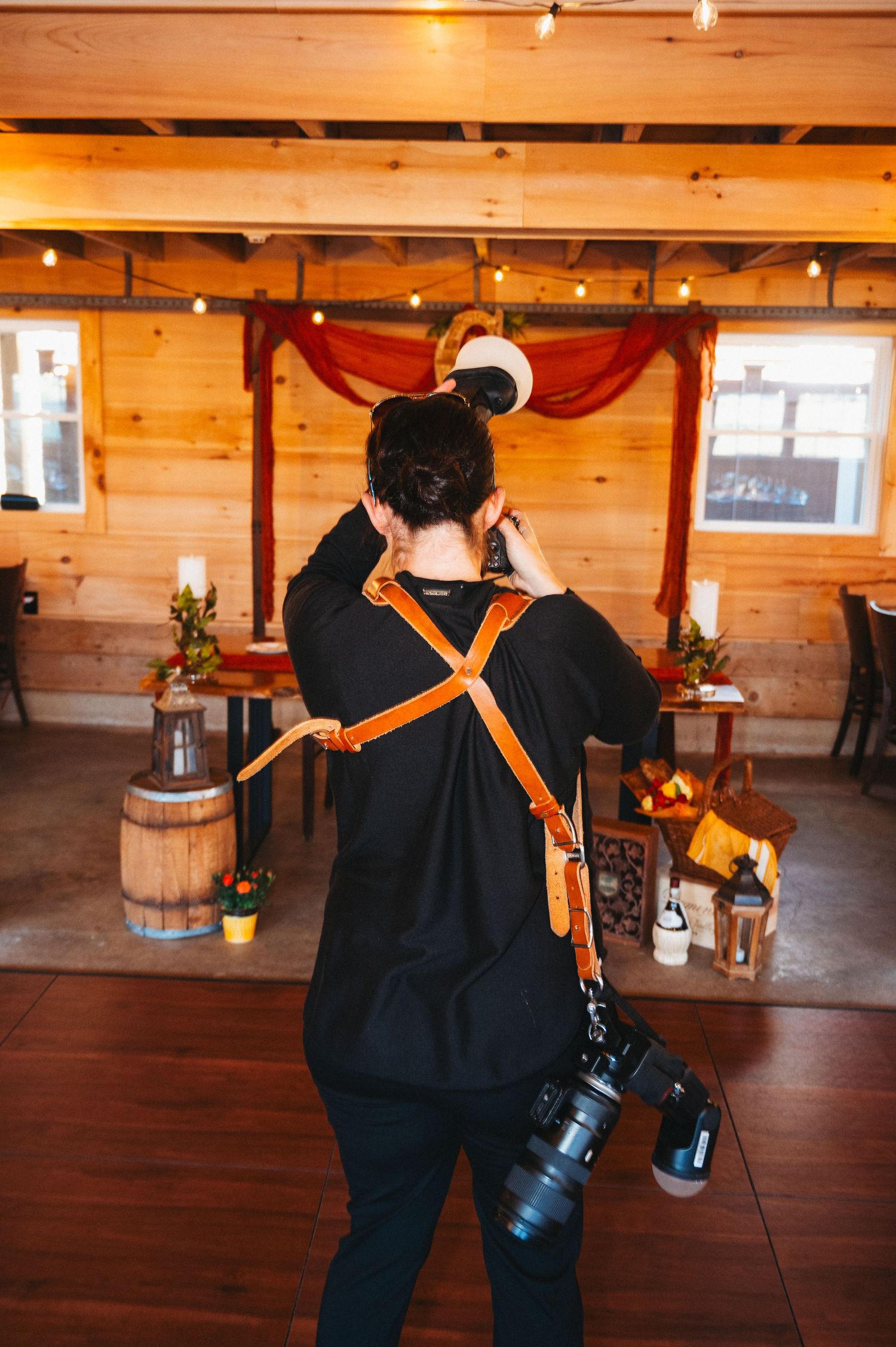 Photographer with orange camera strap holding DSLR camera in rustic wood-paneled venue with string lights.