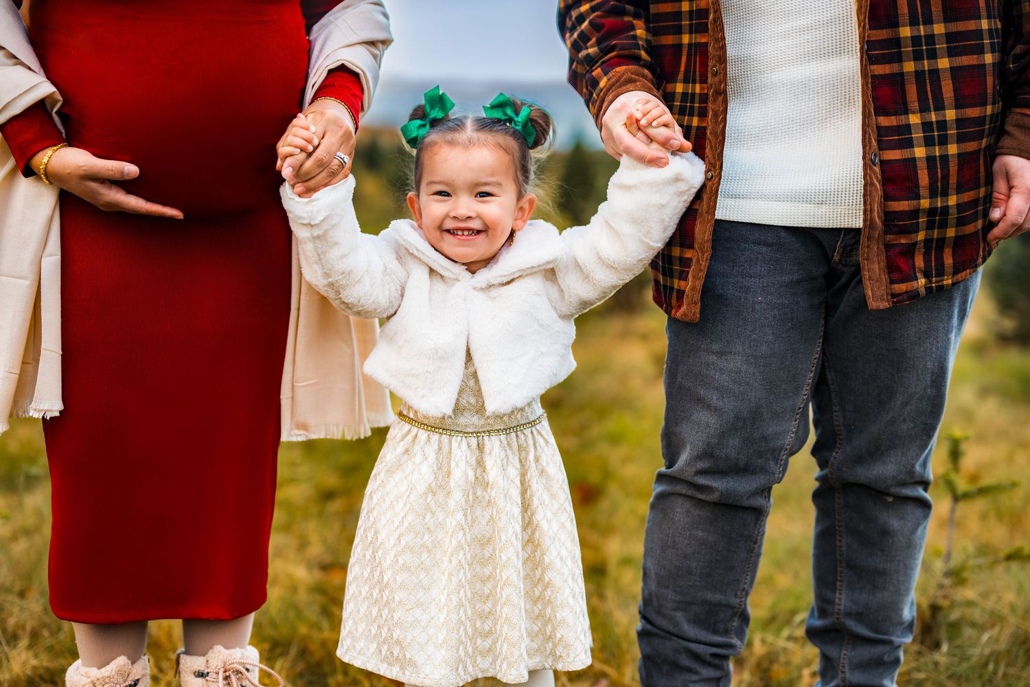 Happy toddler girl in white dress holds parents hands outdoors during family maternity photo session.