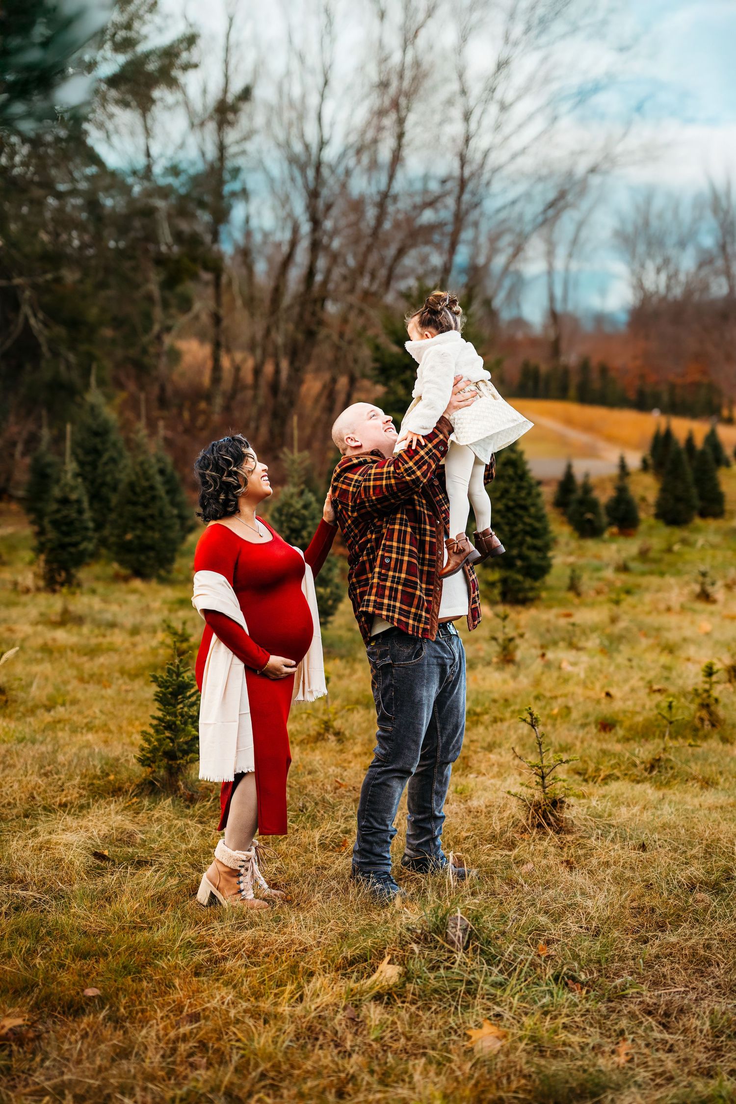 Pregnant woman in red dress with man lifting toddler at Christmas tree farm in autumn.