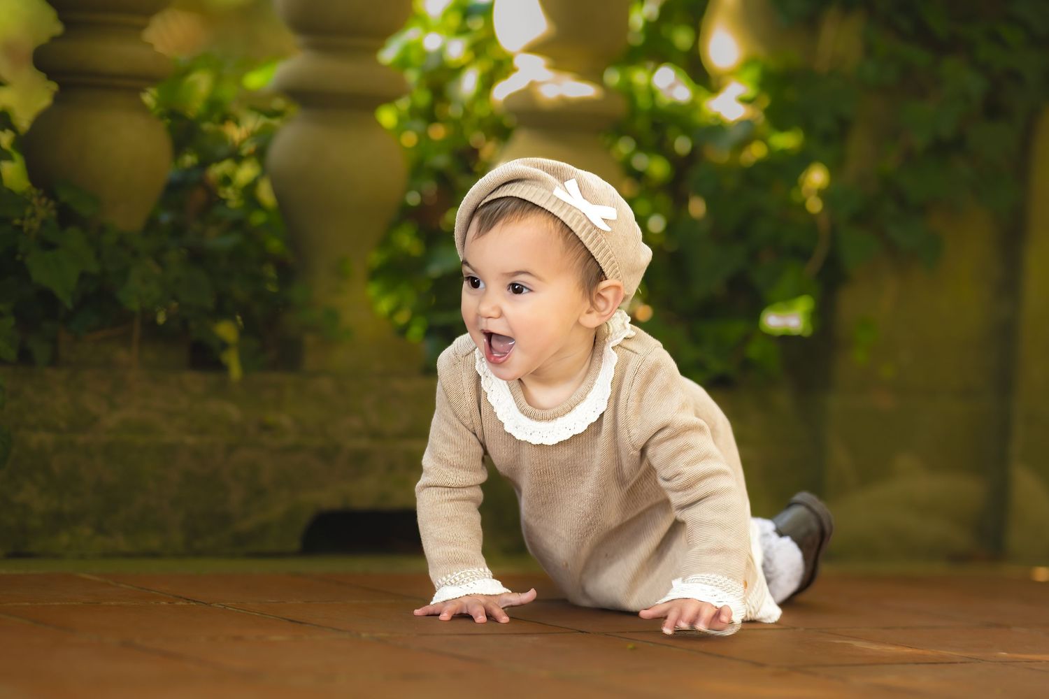 Adorable baby in beige outfit and hat crawling on wooden floor with green plants in background.