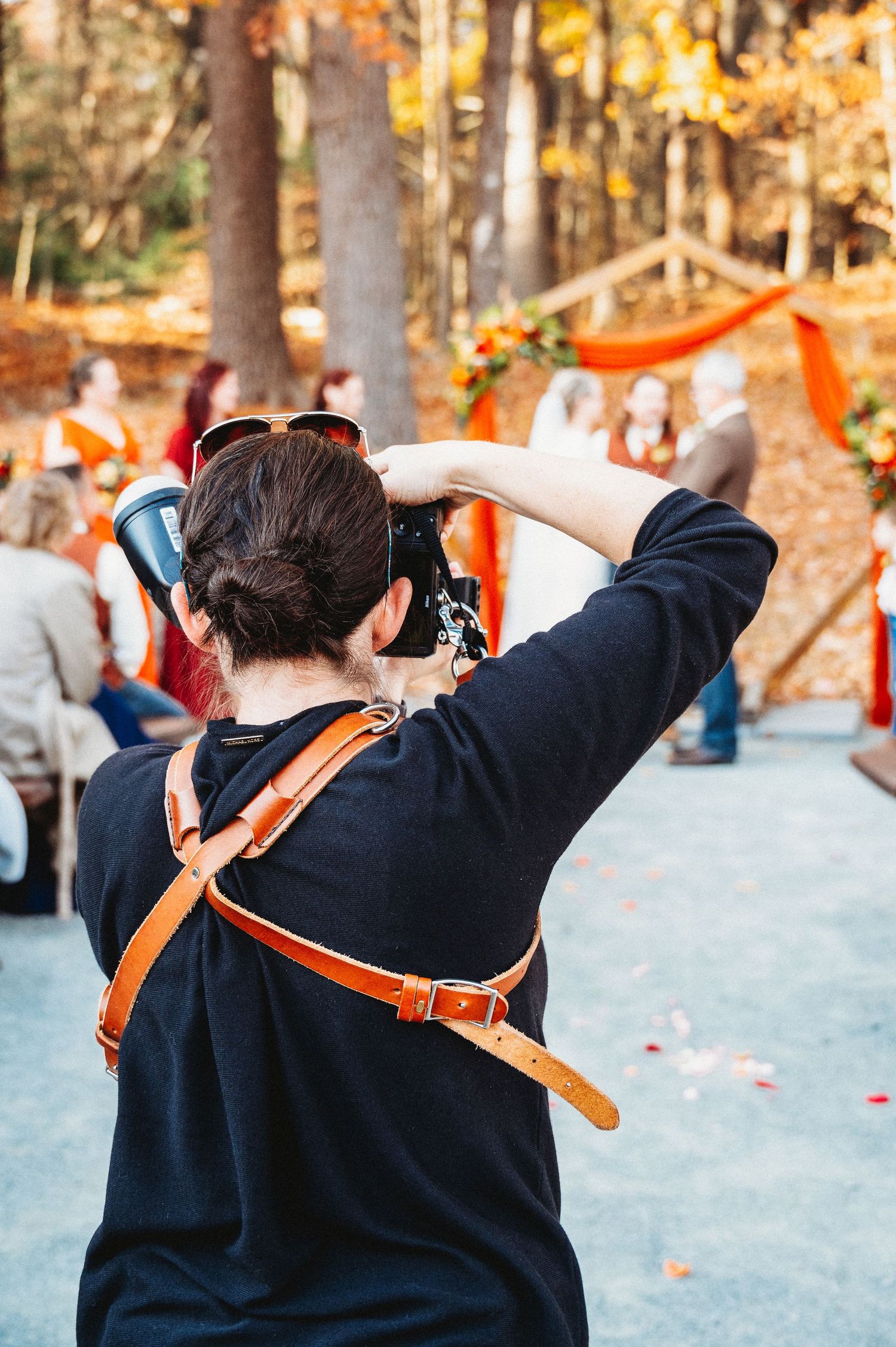 Photographer capturing an outdoor fall wedding ceremony in the woods with orange floral decorations.