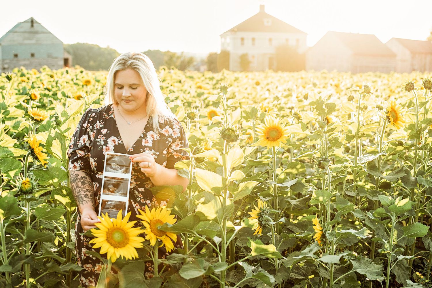 Pregnant woman holding ultrasound photo in sunny sunflower field wearing floral dress for maternity announcement.