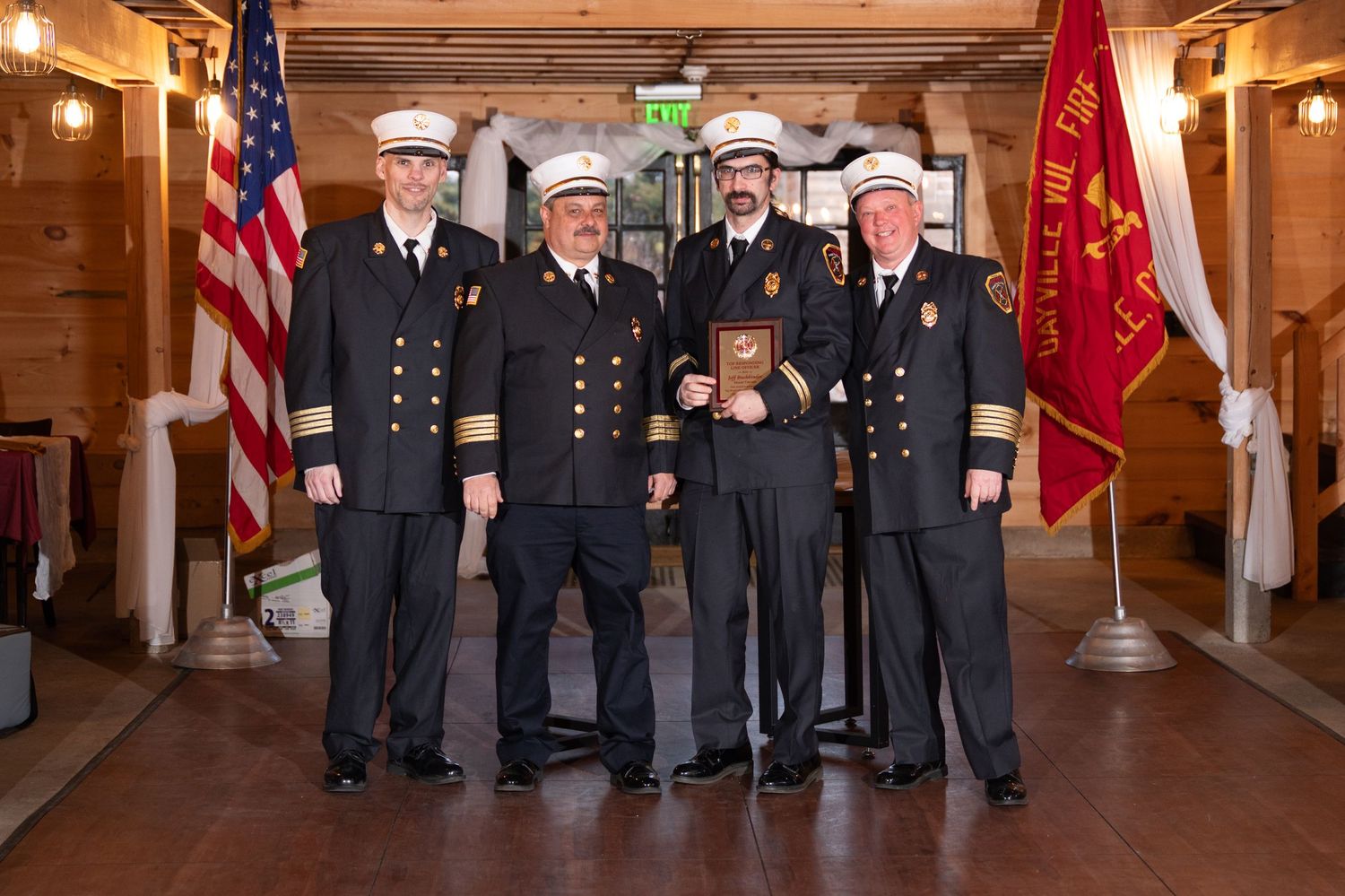 Five firefighters in dress uniforms pose formally at an awards ceremony, with one holding a plaque.